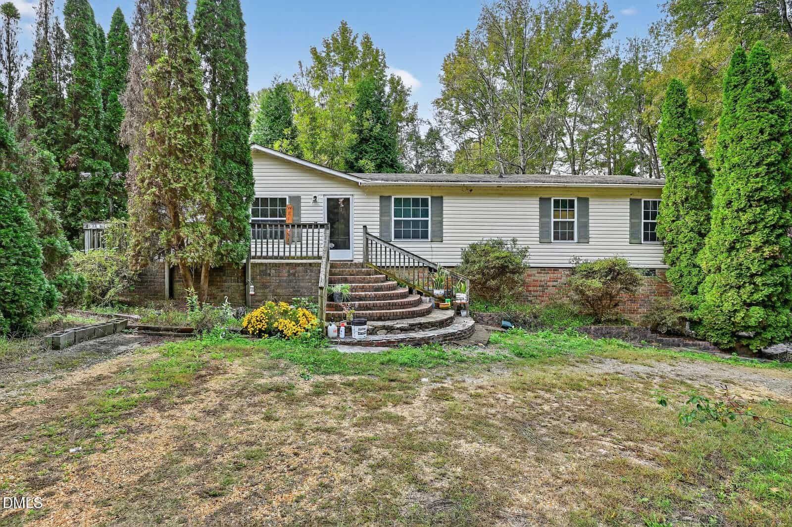 Charming single-story house at 4 Park Ave, Franklinton, with light siding, a brick foundation, and a welcoming front porch featuring black railings. Curved brick steps lead to the porch, while tall shrubs and trees surround the property. The yard is mostly bare with patches of grass and plants, adding to its tranquil appeal.