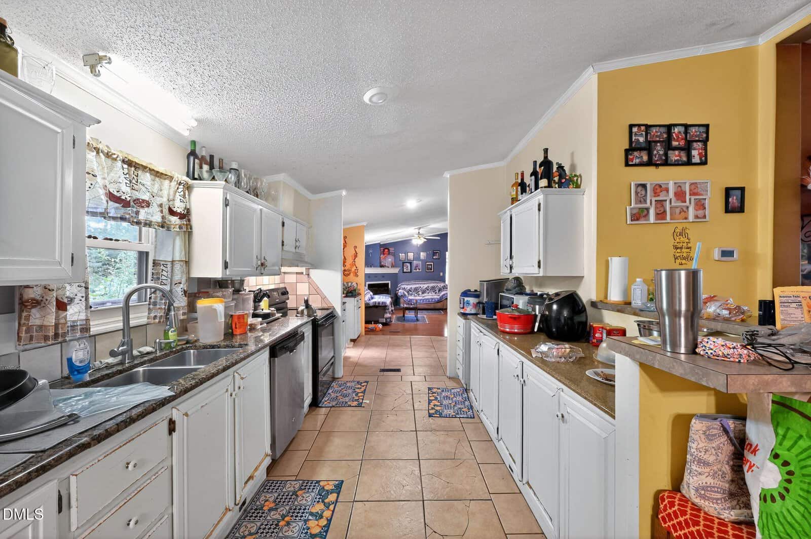 A lived-in kitchen with white cabinets, granite counters, and tile floor. Small appliances, dishes, and food items are on the counters. The kitchen opens to a living room with a couch and TV visible in the background.