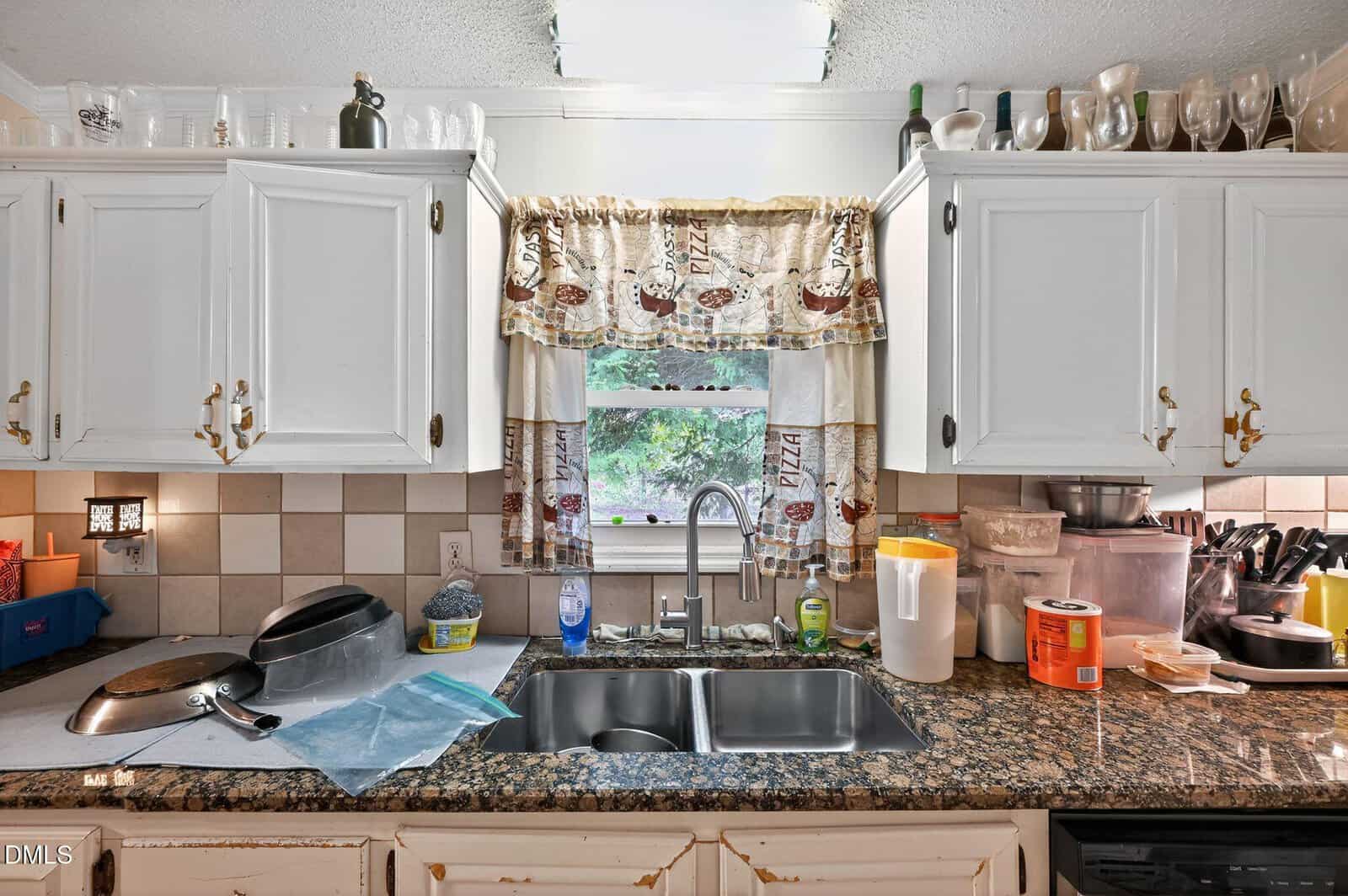 Kitchen with white cabinets, granite countertops, and a double sink beneath a window with a pizza-themed curtain. Counter has various containers, a dish rack, cleaning supplies, and kitchen items. Shelves above hold glasses and bottles.