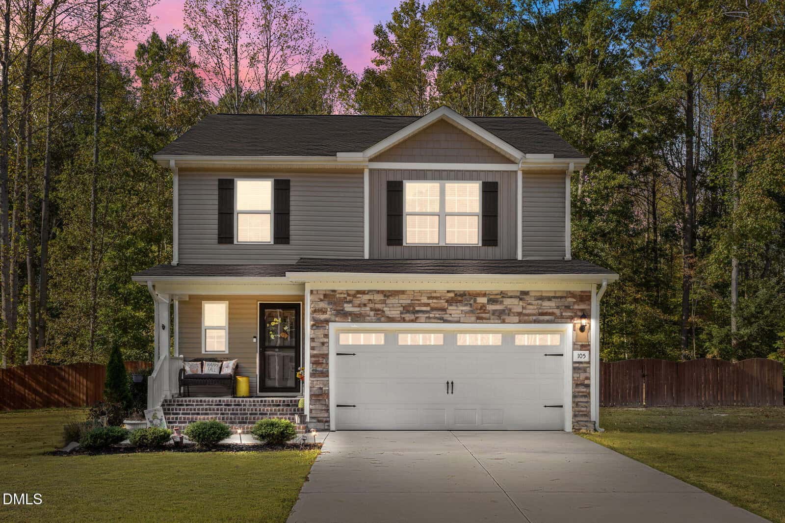 Two-story suburban house with a double garage, front porch, stone and siding exterior, and a landscaped yard at 105 Teal Drive in Youngsville, set against a background of tall trees at sunset.