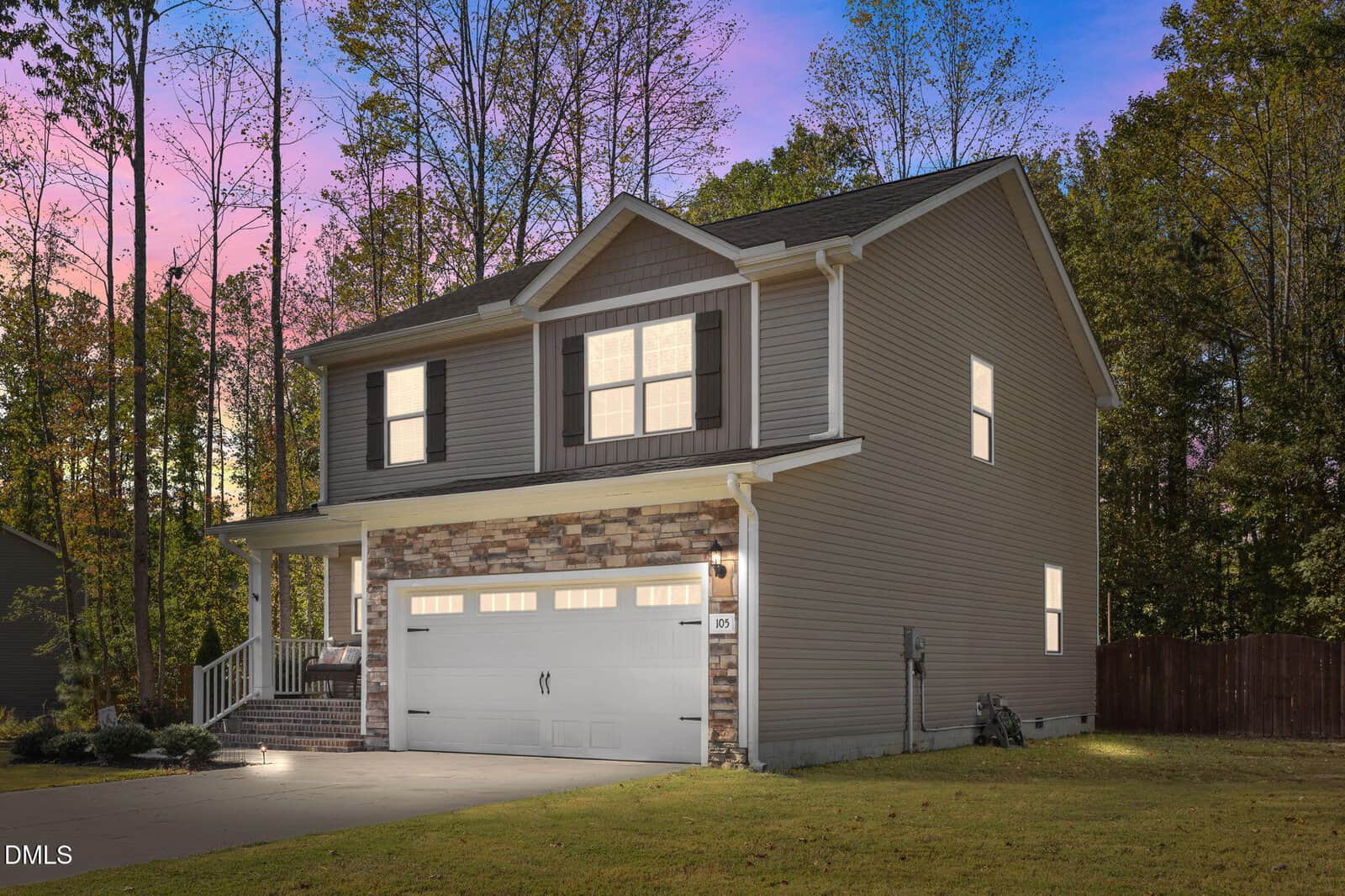 Two-story house with beige siding, stone accents on the lower front, white garage door, front porch, and lit windows. Surrounded by tall trees, the sky shows a pink and blue gradient at dusk. Concrete driveway in foreground.