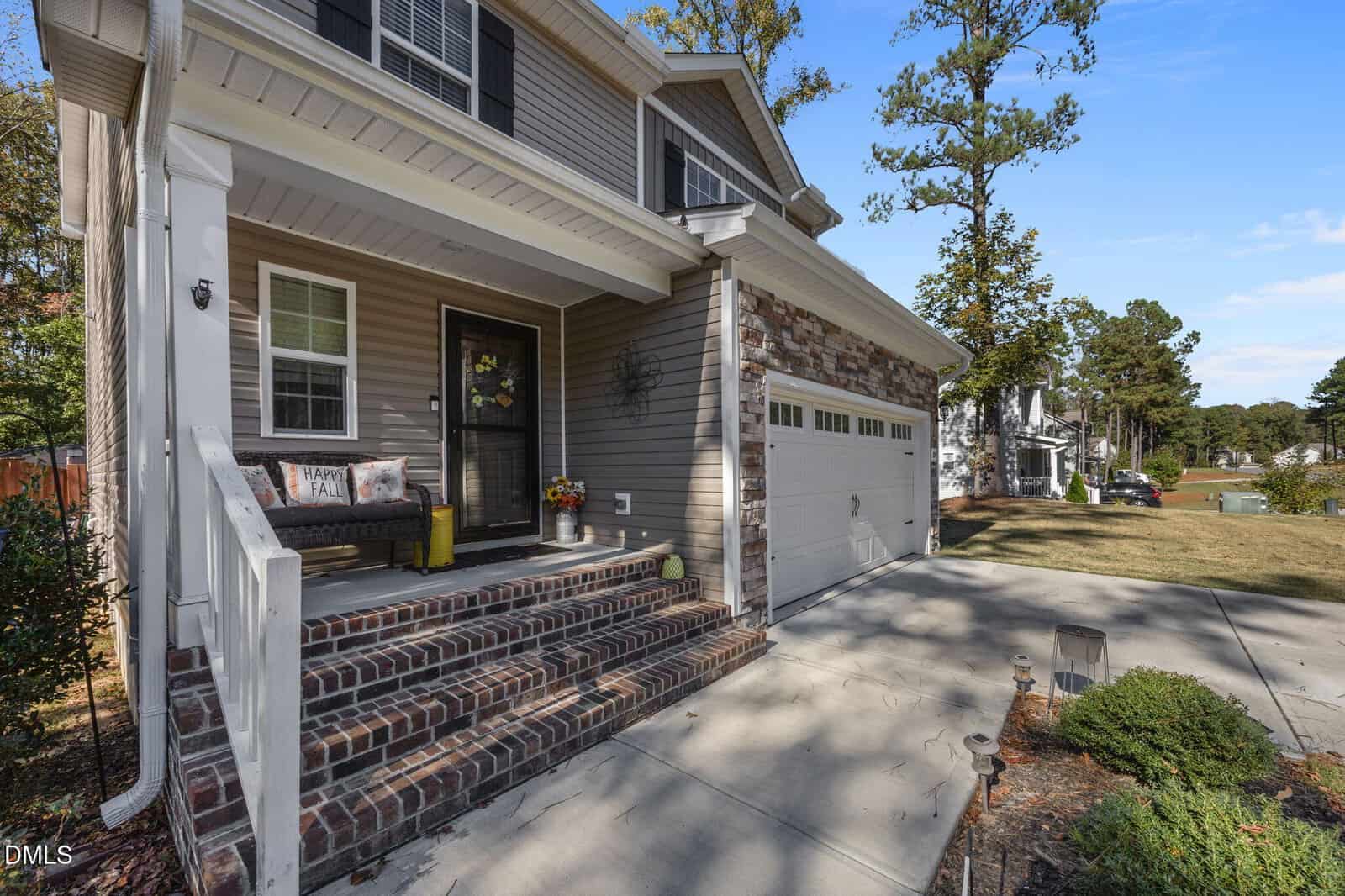 Two-story suburban house with beige siding, stone accents, and a white garage. The front porch has brick steps, a bench, potted plants, and a “Happy Fall” sign. Trees and neighboring houses are visible in the background.