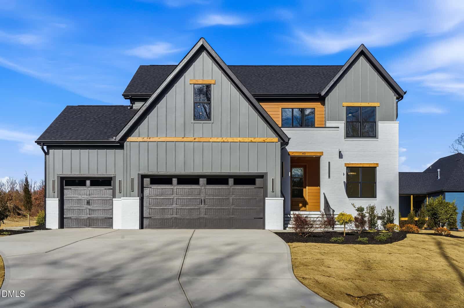 A modern two-story house with gray vertical siding, white brick, black-framed windows, and three dark garage doors. The home features natural wood accents and is surrounded by a newly landscaped yard.