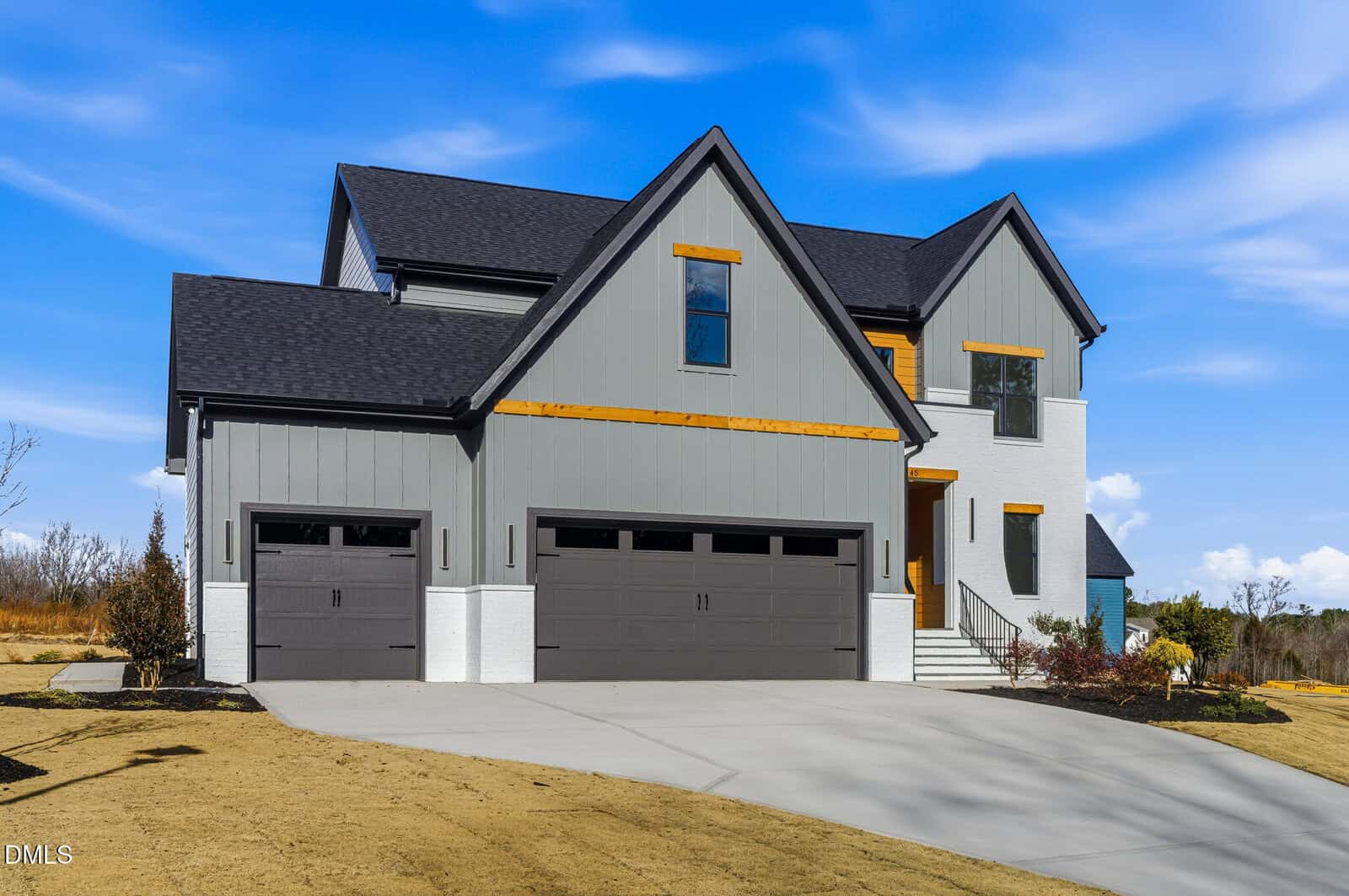Two-story modern house with gray vertical siding, white brick lower exterior, three-car garage, black roof, and wood trim accents, set on a lawn with a curved driveway under a blue sky with clouds.