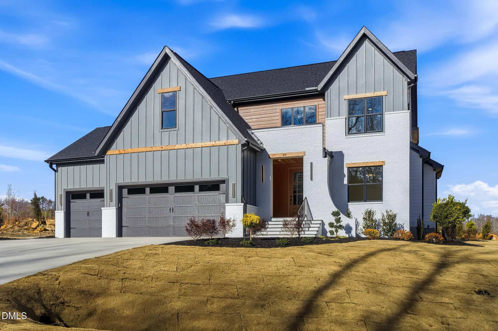 A modern two-story house with gray siding, black roof, and three-car garage. The front yard has newly laid sod and small shrubs, with a concrete driveway and steps leading to the entrance. The sky is clear and blue.
