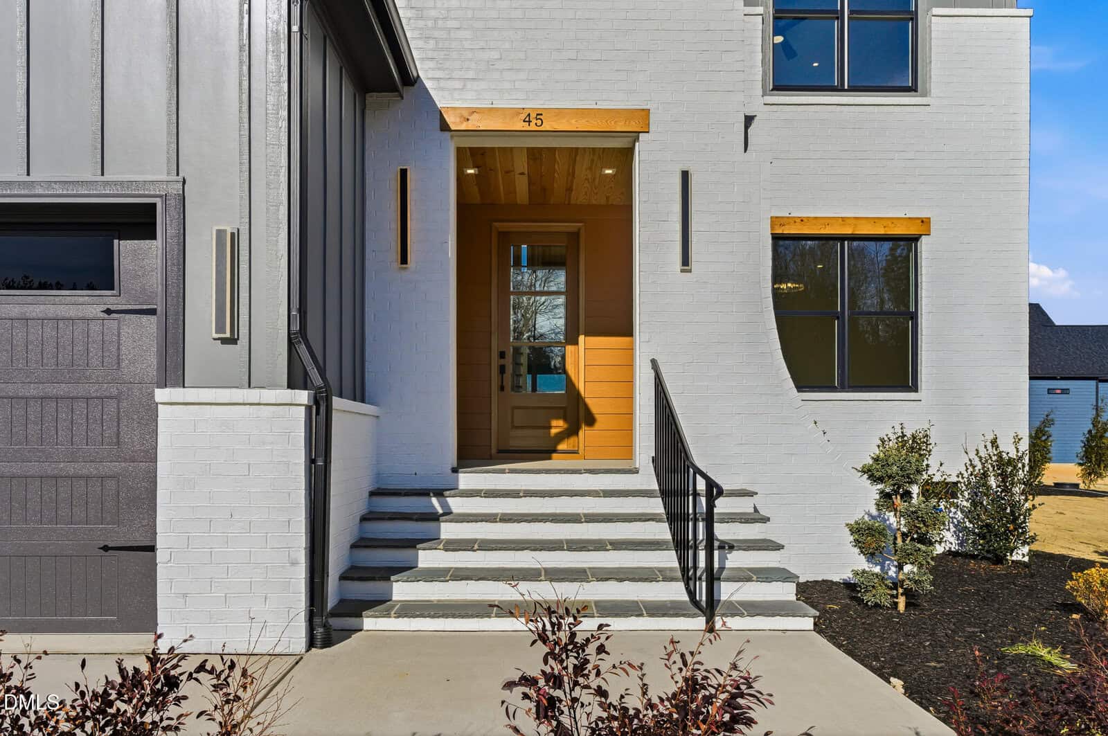 A modern house entrance with light-colored brick, a wooden door, and a small porch with steps and black railings. There are large windows, a garage door to the left, and landscaping with shrubs in front.