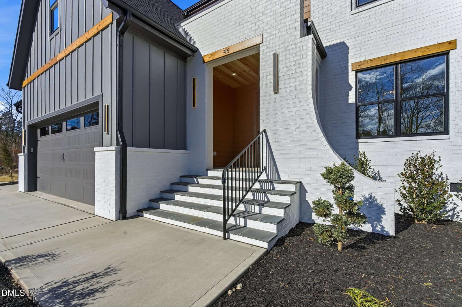Modern house exterior with white brick and gray siding, a two-car garage on the left, a short stairway with a metal railing leading to a recessed front entry, and small shrubs planted along the front walkway.