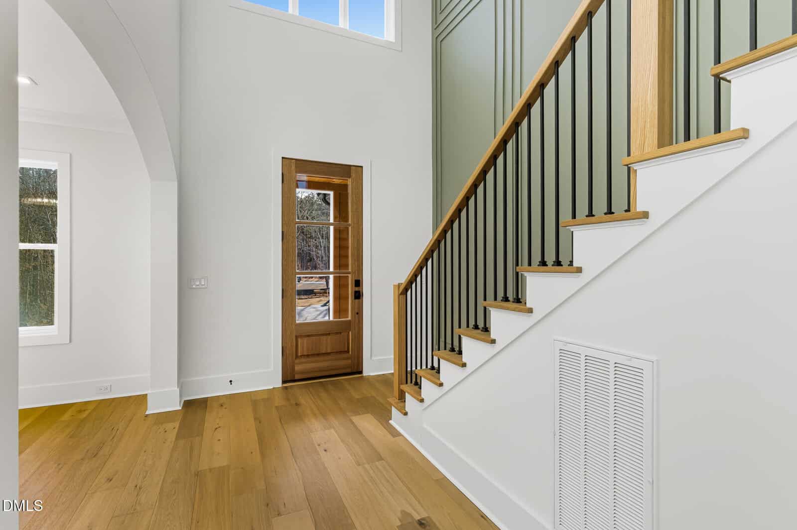 A bright foyer with light wood floors, a wooden front door with glass, a staircase with wood treads and black spindles, and white walls with tall windows.