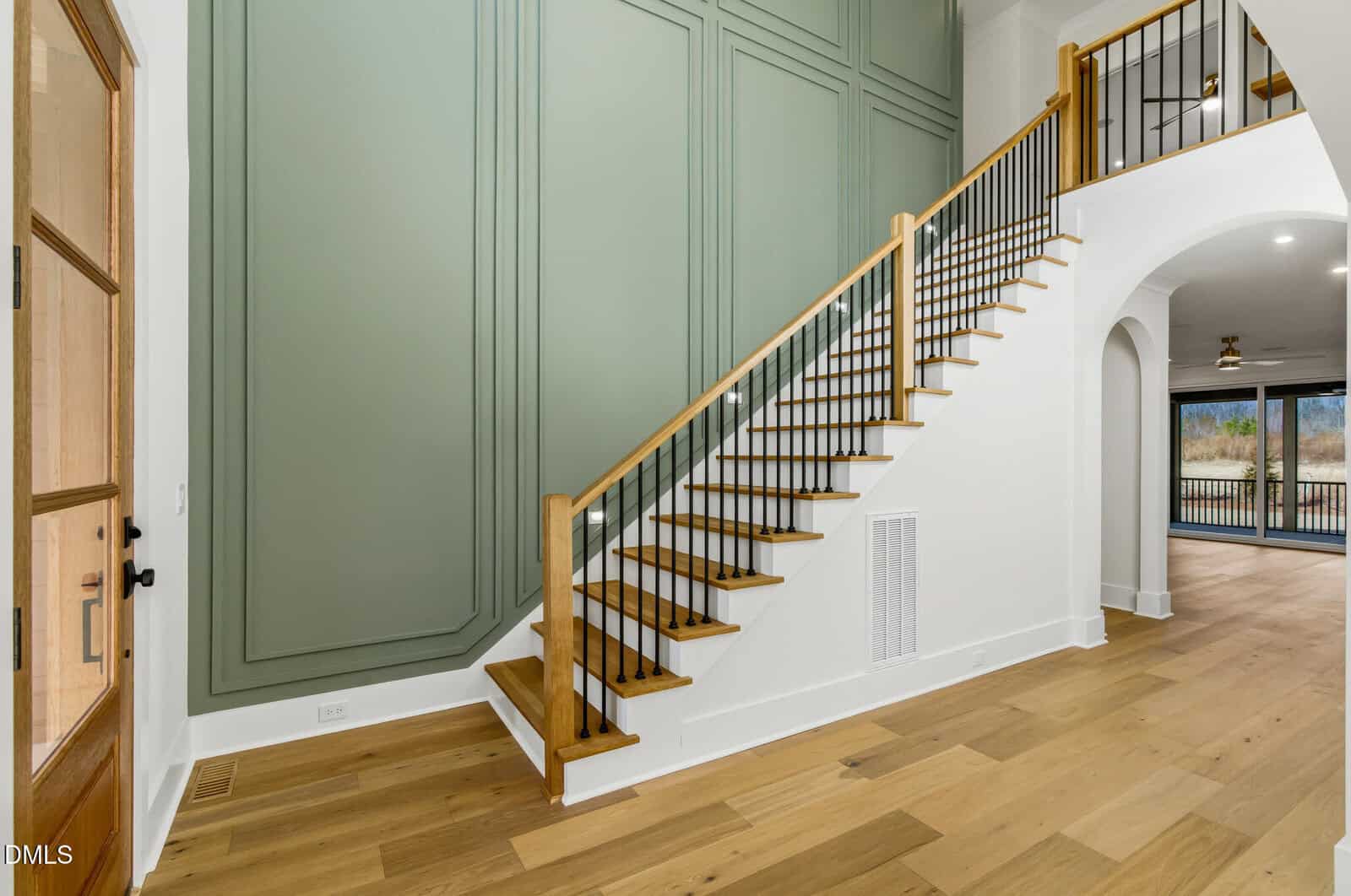 Modern entryway with light wood floors, a staircase with wooden handrail and black balusters, large green accent wall, white walls, and an open doorway leading to another room with large windows.