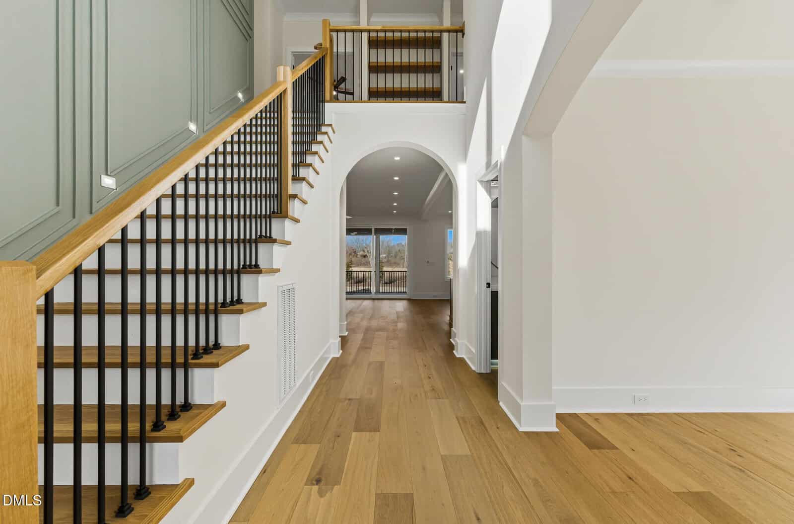 Modern home interior with light wood floors, a staircase with black railings, green wall paneling, and an open hallway leading to large windows and a bright outdoor view.