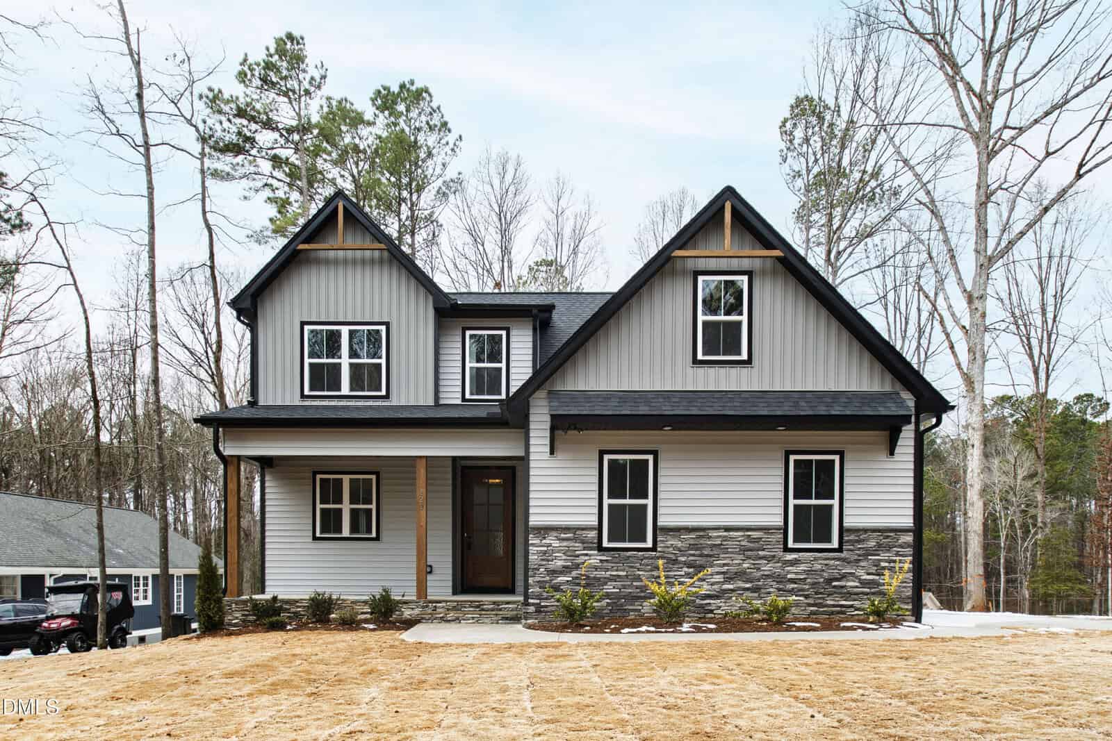 A modern two-story house with gray siding, stone accents, black trim, and a covered front porch. The yard is freshly seeded, and the house is surrounded by trees. Another house is visible to the left.