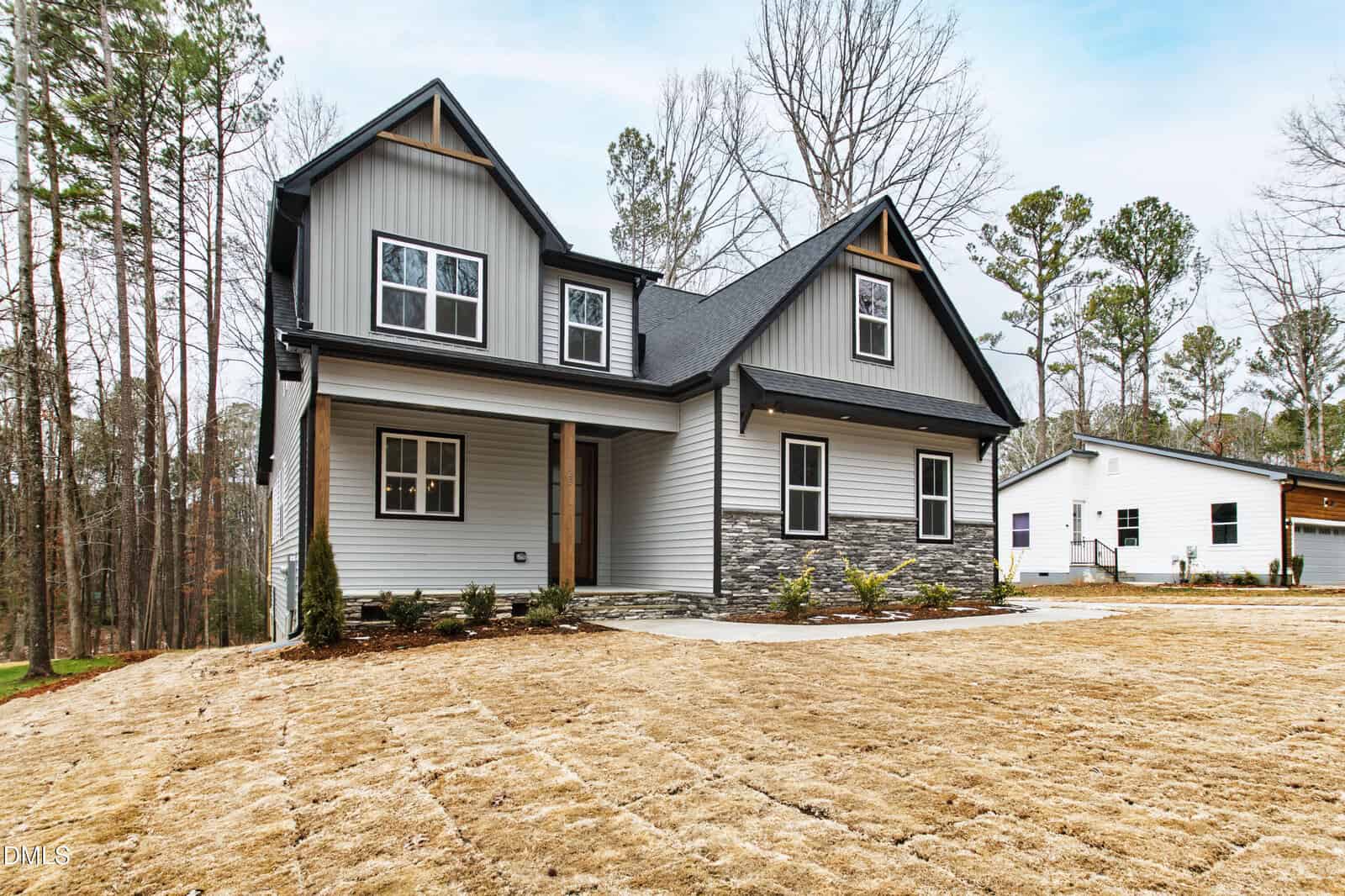 A modern two-story house with gray siding, black roof, stone accent, and a covered front porch, set on a newly seeded lawn with young landscaping and surrounded by trees.