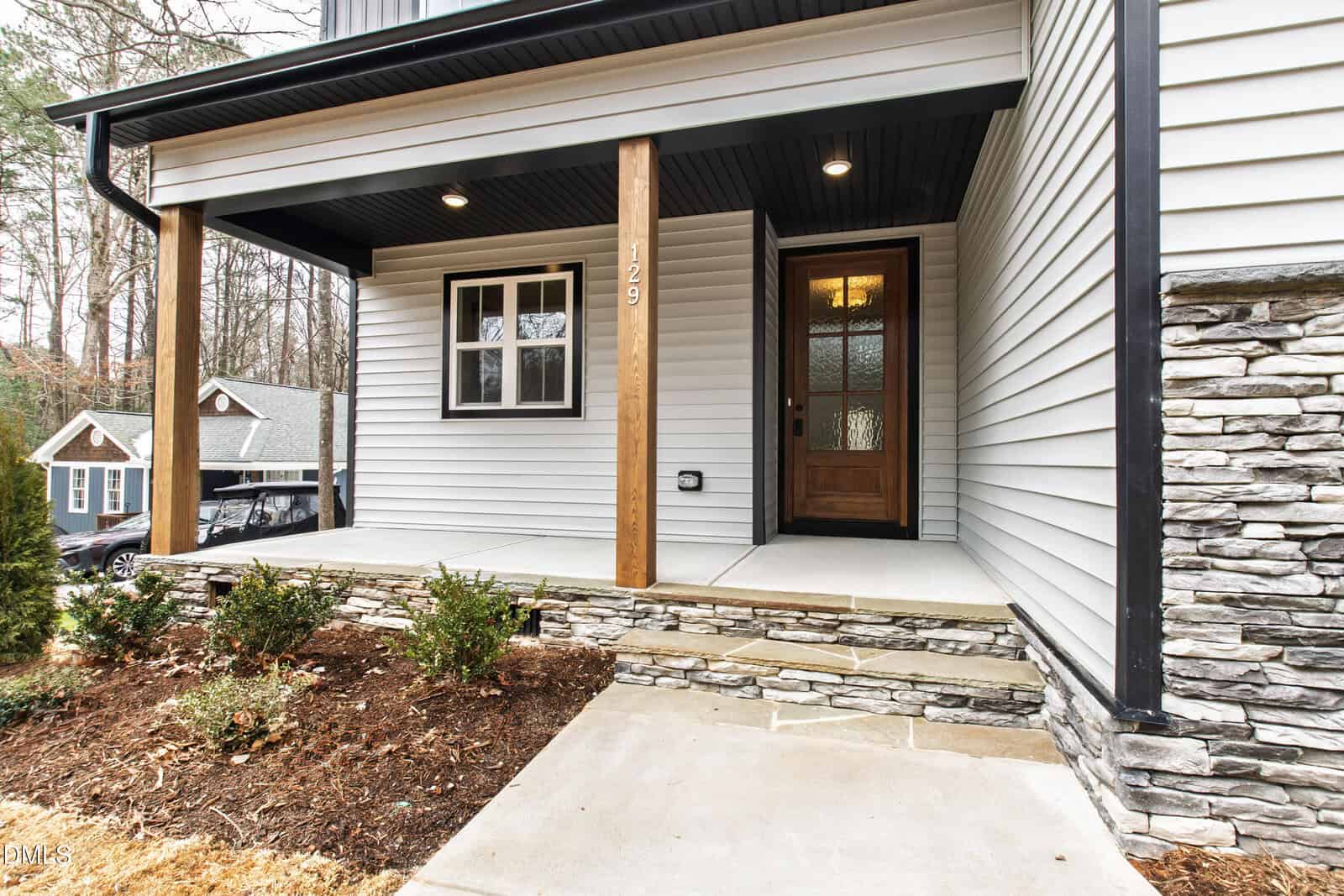 A modern house front porch with white siding, stone accents, wooden posts, a wooden door with glass, and small shrubs planted in a mulched bed. Steps lead up to the porch from a concrete walkway.