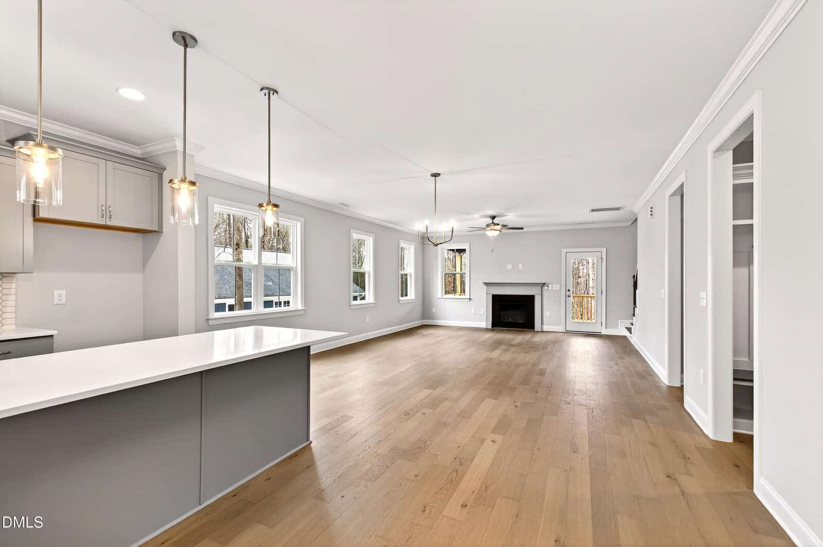 A spacious open-concept living area with light wood floors, large windows, modern pendant lights over a kitchen island, gray cabinets, and a white fireplace in the living room area. Walls are painted light gray.