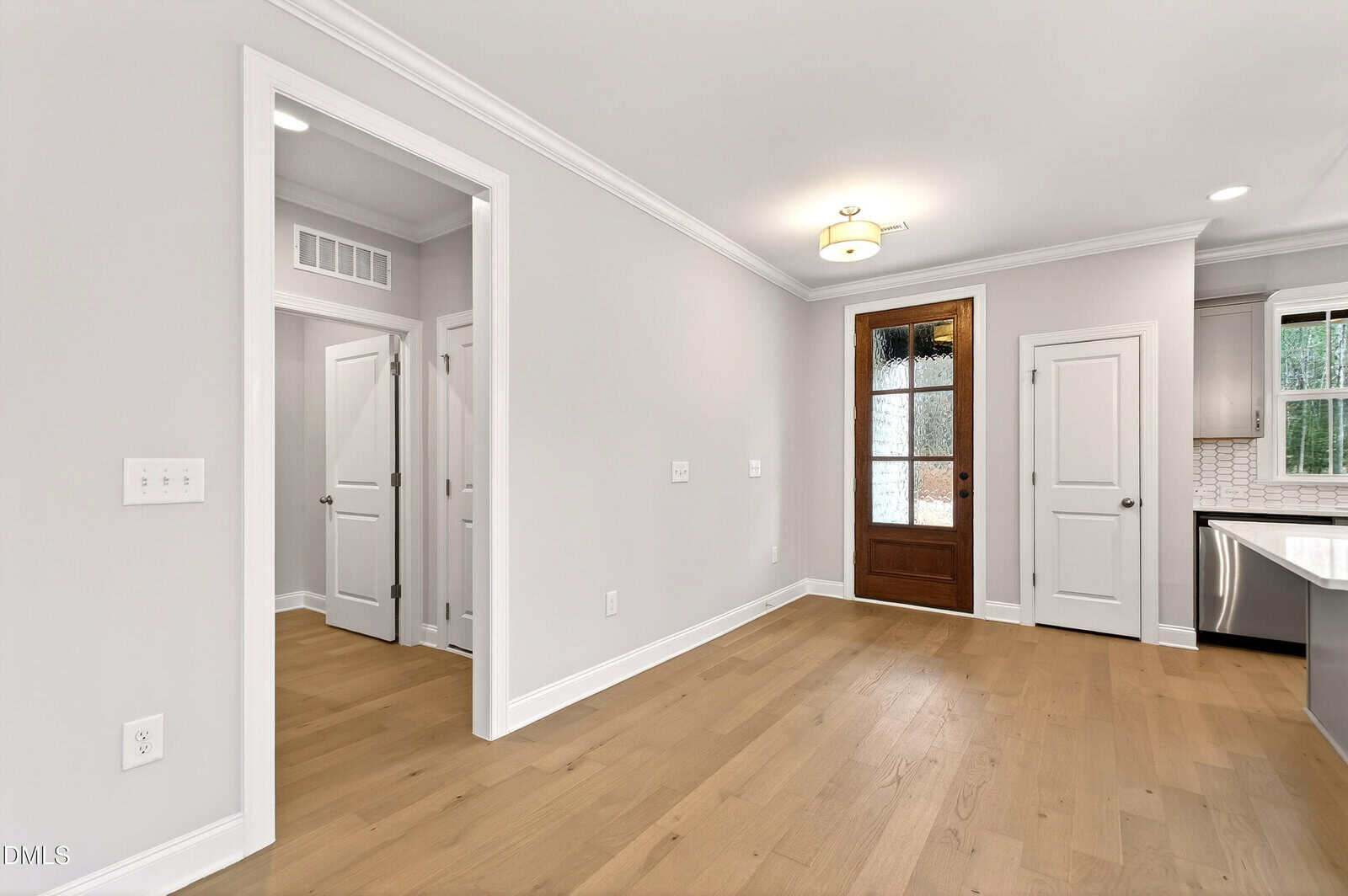 A bright, empty room with light wood flooring, white walls, and white trim. There are several white doors, a wooden exterior door with glass panes, and a partial view of a modern kitchen with tile backsplash.