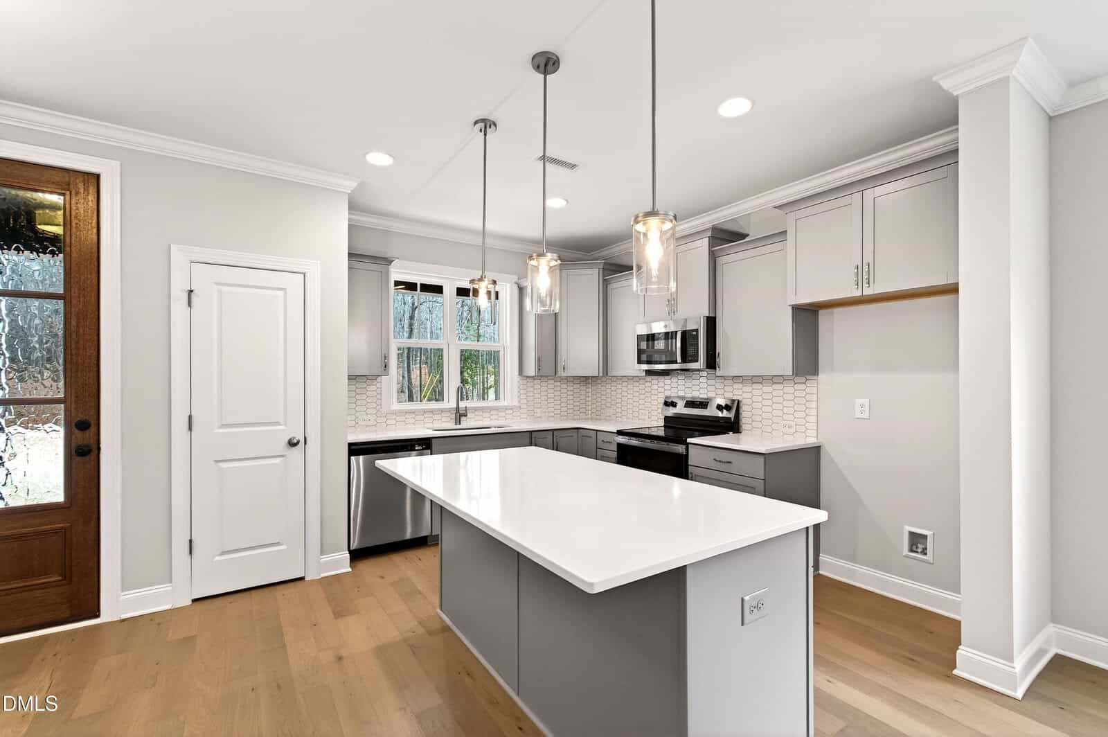 Modern kitchen with light gray cabinets, white countertops, stainless steel appliances, a center island with pendant lights, hardwood floors, and a large window above the sink. A wooden door is visible on the left.