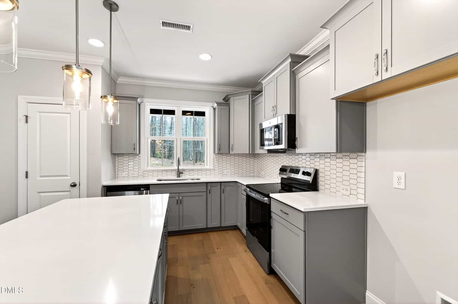 Modern kitchen with light gray cabinets, white countertops, stainless steel appliances, pendant lights, a window above the sink, light wood flooring, and a white tile backsplash.