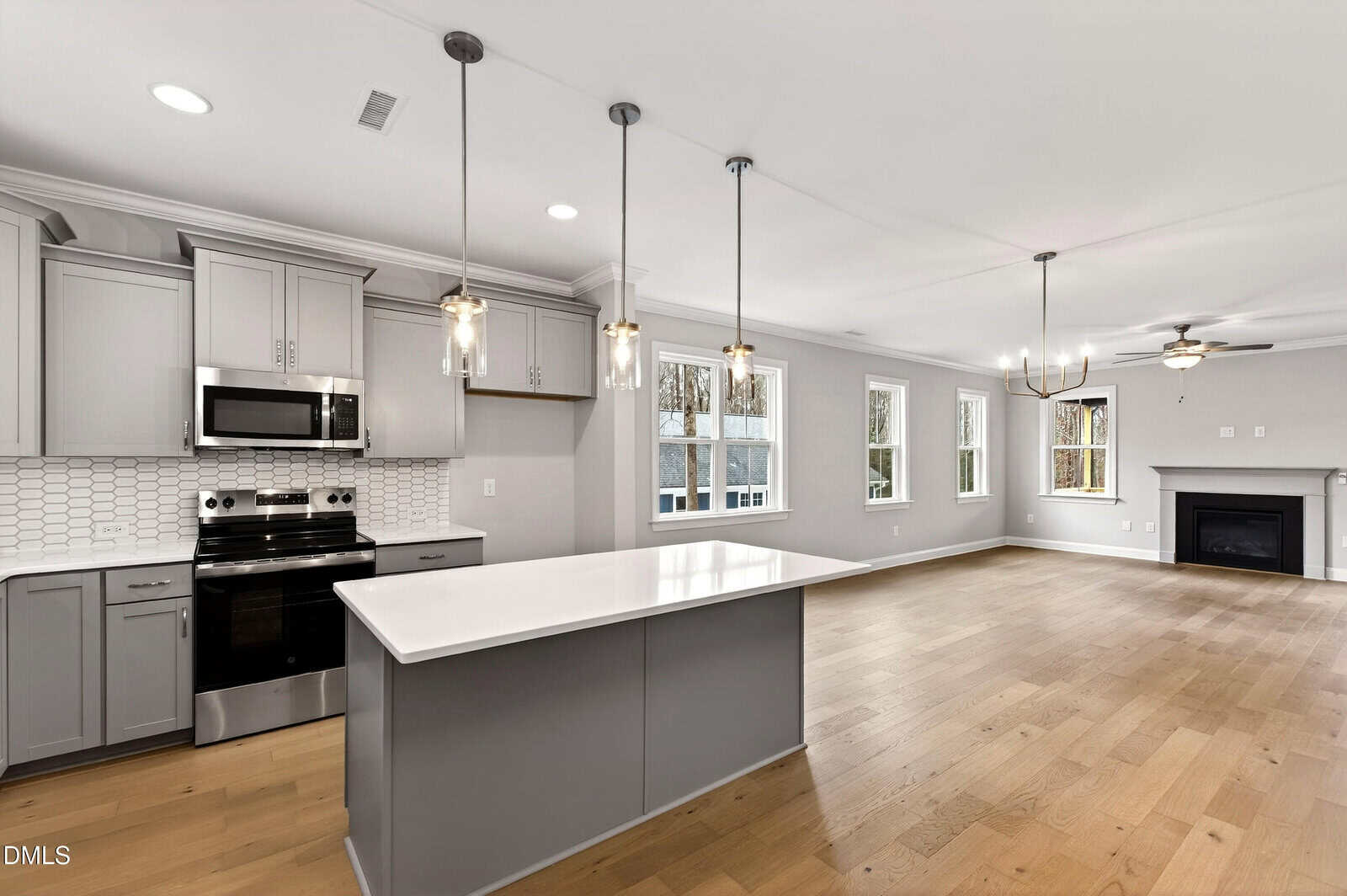 Modern, open-concept kitchen and living area with light wood flooring, gray cabinets, stainless steel appliances, pendant lighting, a large white island, and multiple windows providing natural light.