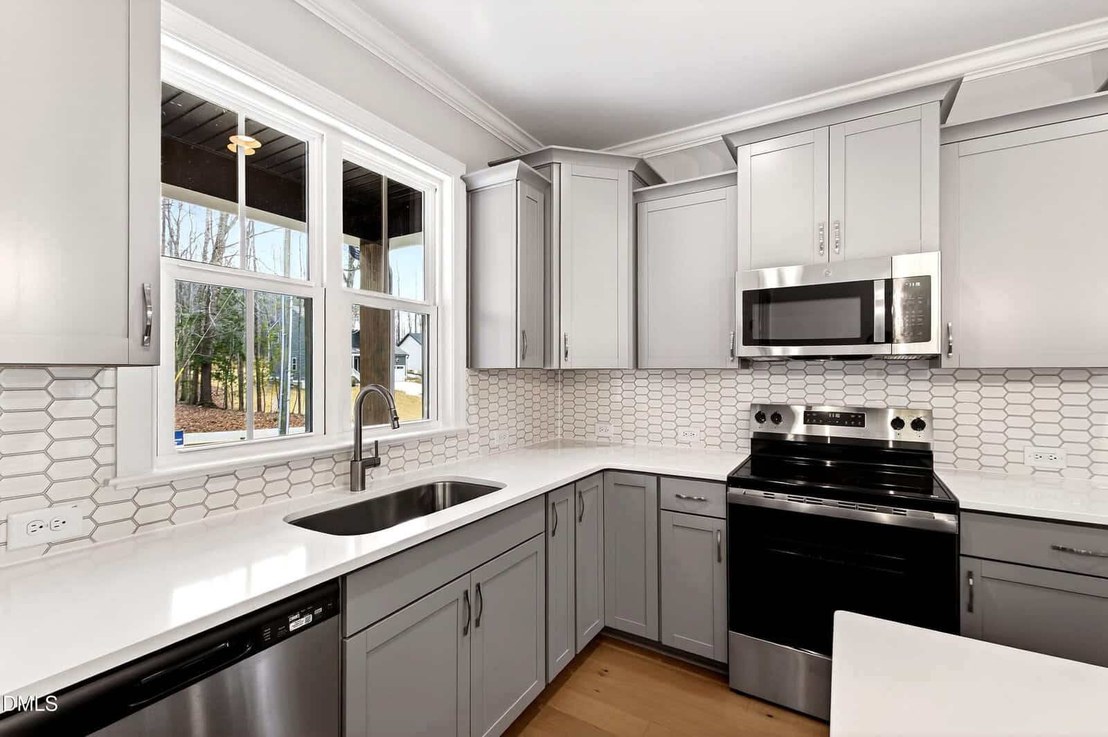 Modern kitchen with gray cabinets, white countertops, stainless steel appliances, a large window over the sink, and a white hexagonal tile backsplash. Light wood flooring and outdoor view through the window.