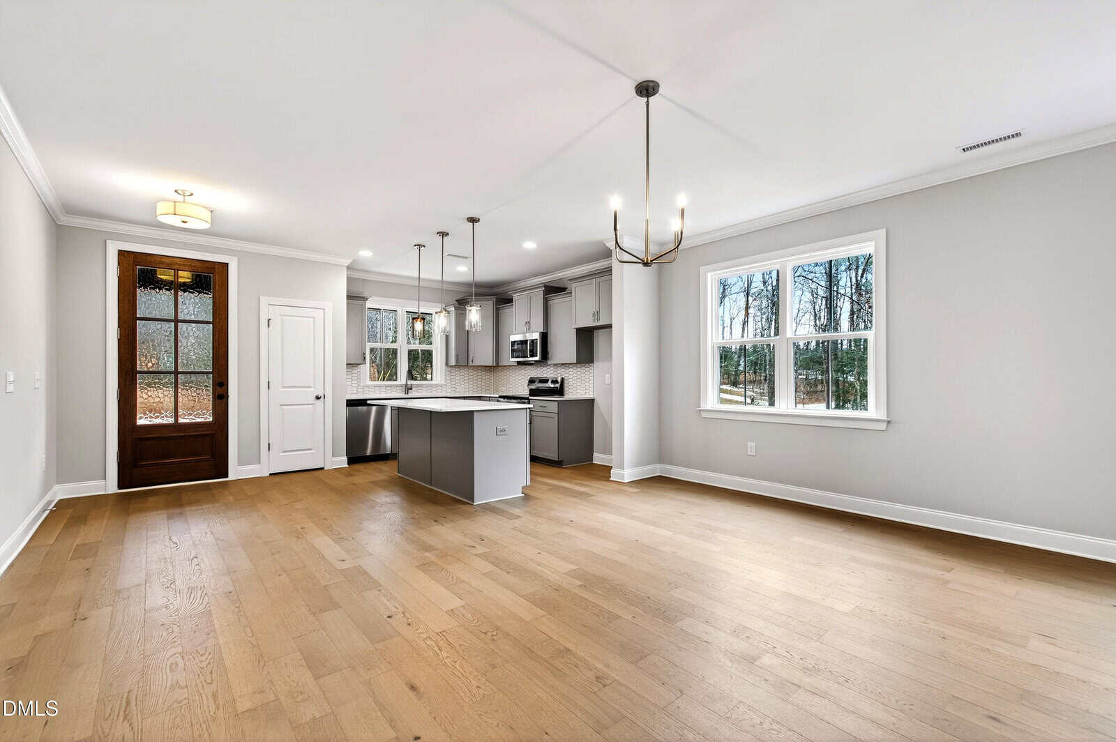 Modern kitchen and dining area with light wood floors, gray island, stainless steel appliances, white cabinets, pendant lighting, and large windows letting in natural light; wooden front door visible in the background.
