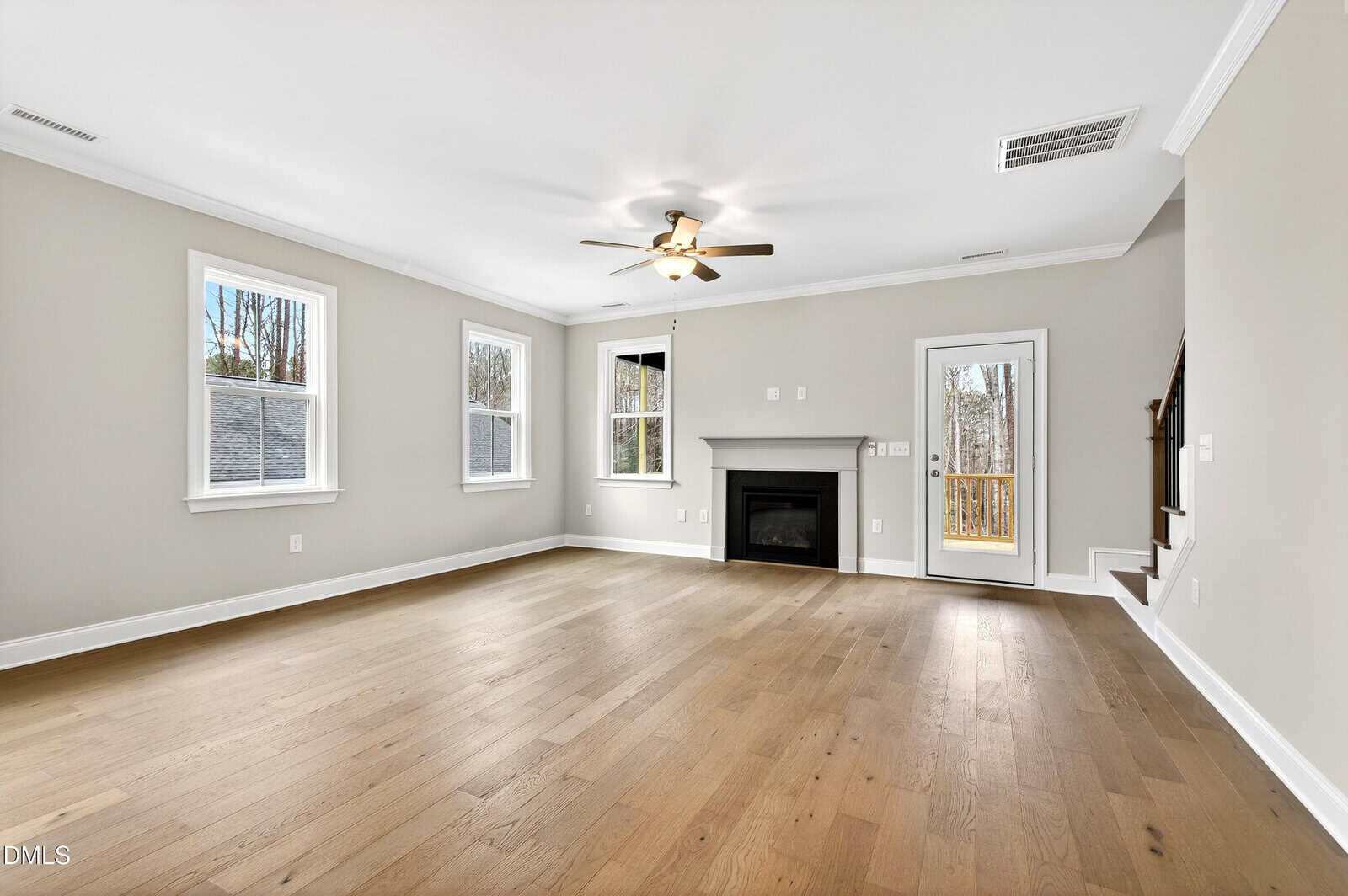 Spacious, empty living room with light wood floor, beige walls, three windows, a ceiling fan, fireplace, and a door next to a staircase.