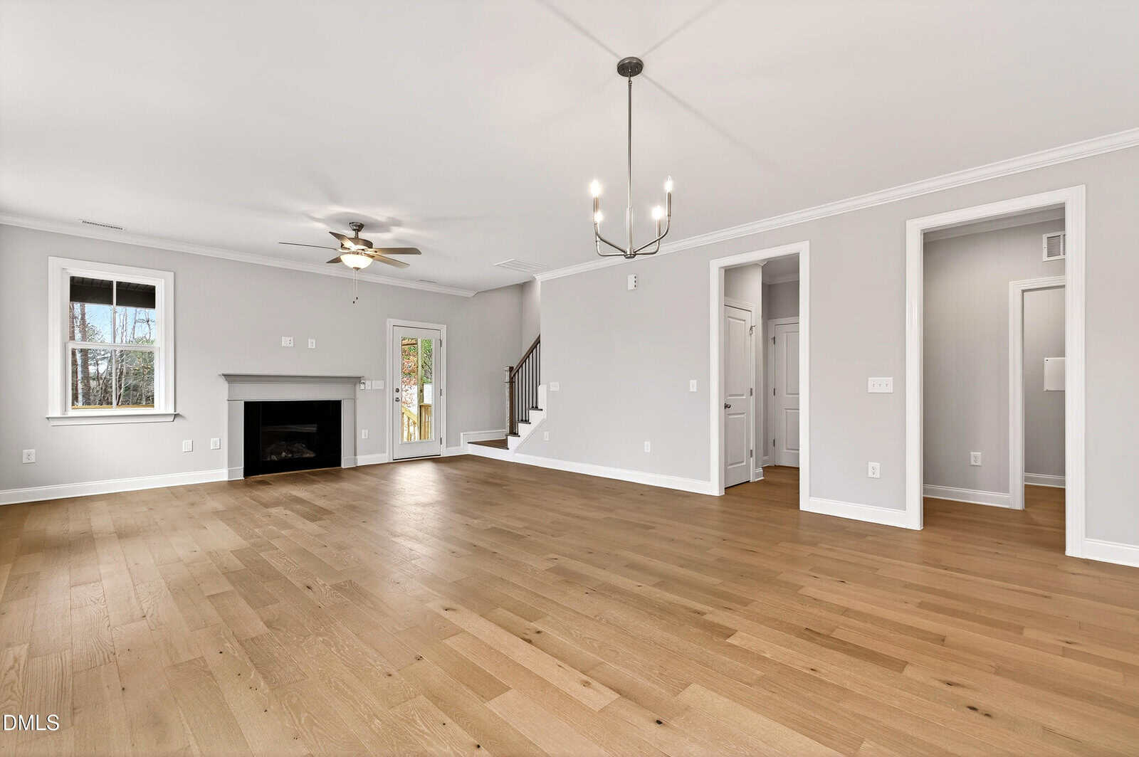 Spacious, empty living room with light wood floors, gray walls, a black fireplace, large windows, a modern chandelier, and an open staircase leading upstairs.