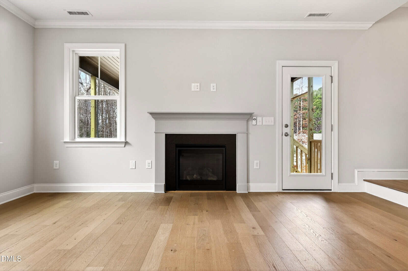 A bright, empty room with light wood flooring, a gray fireplace, white walls, a window on the left, and a glass door on the right that opens to a wooden deck with trees visible outside.