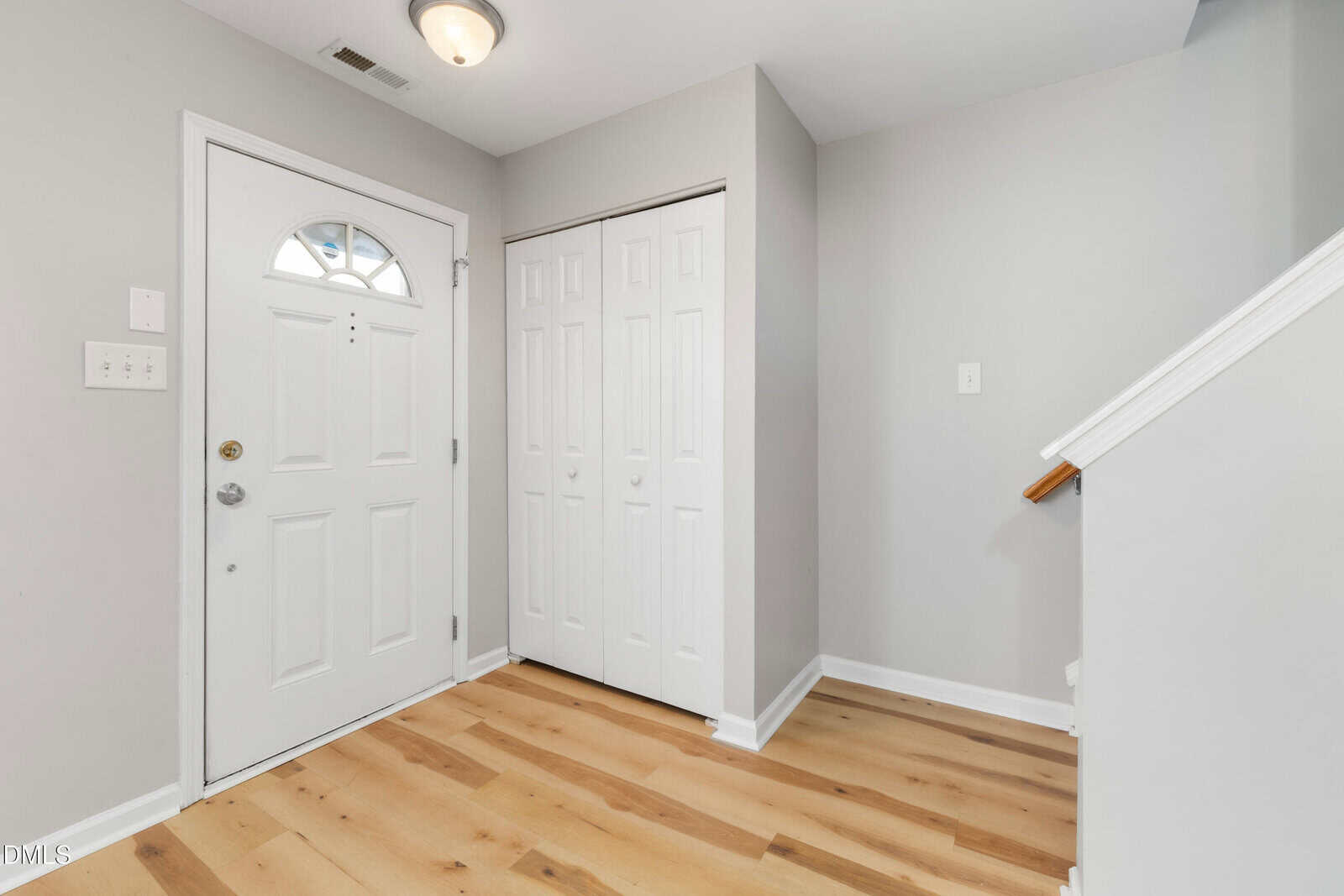 A clean entryway with light wood flooring, a white front door with a window, a white double closet door, light gray walls, and a staircase with a wooden handrail.