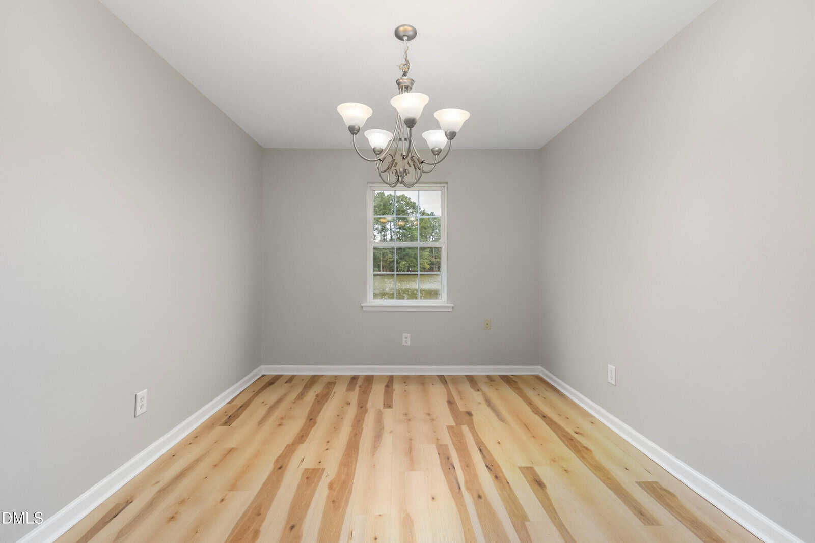 Empty room with light wood flooring, light gray walls, a central window showing greenery outside, and a ceiling-mounted chandelier with five lights. There are white baseboards and electrical outlets on the walls.
