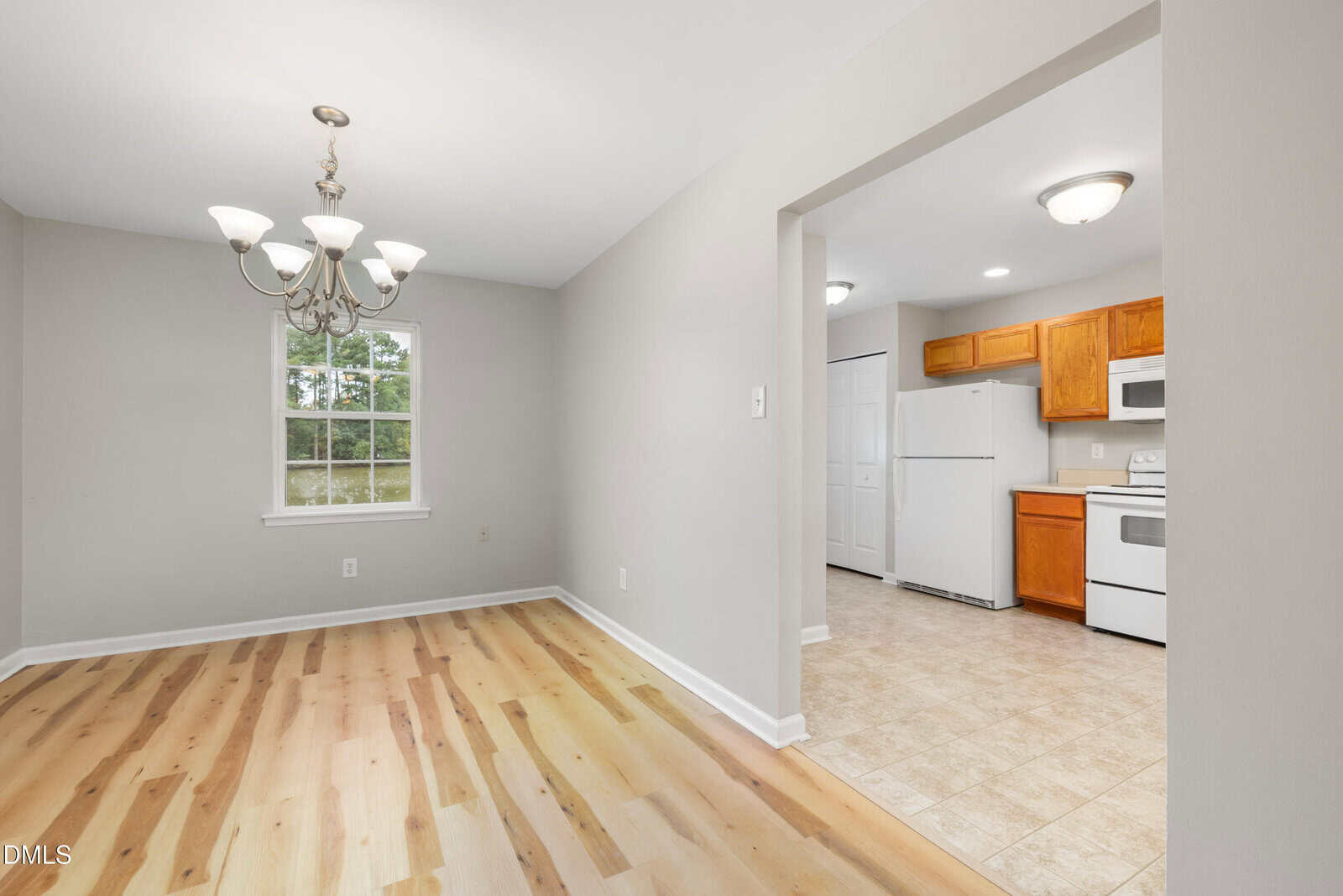 A bright dining area with a chandelier, light wood floors, and a window, adjacent to a kitchen featuring white appliances, wood cabinets, and a tiled floor.