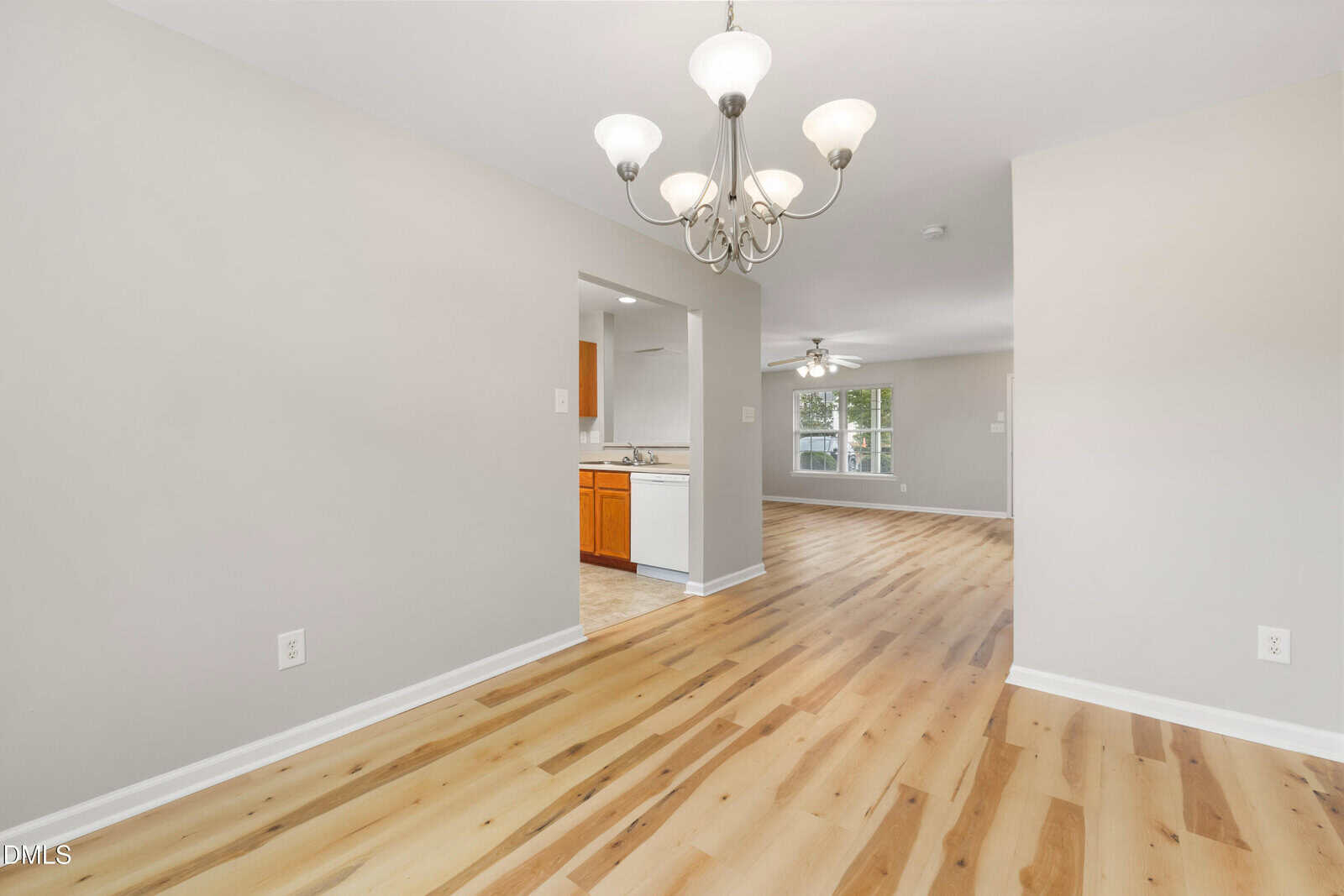 A bright, empty room with light wood floors, beige walls, and a chandelier. An open doorway leads to a kitchen with wood cabinets and another room with a ceiling fan and large windows.