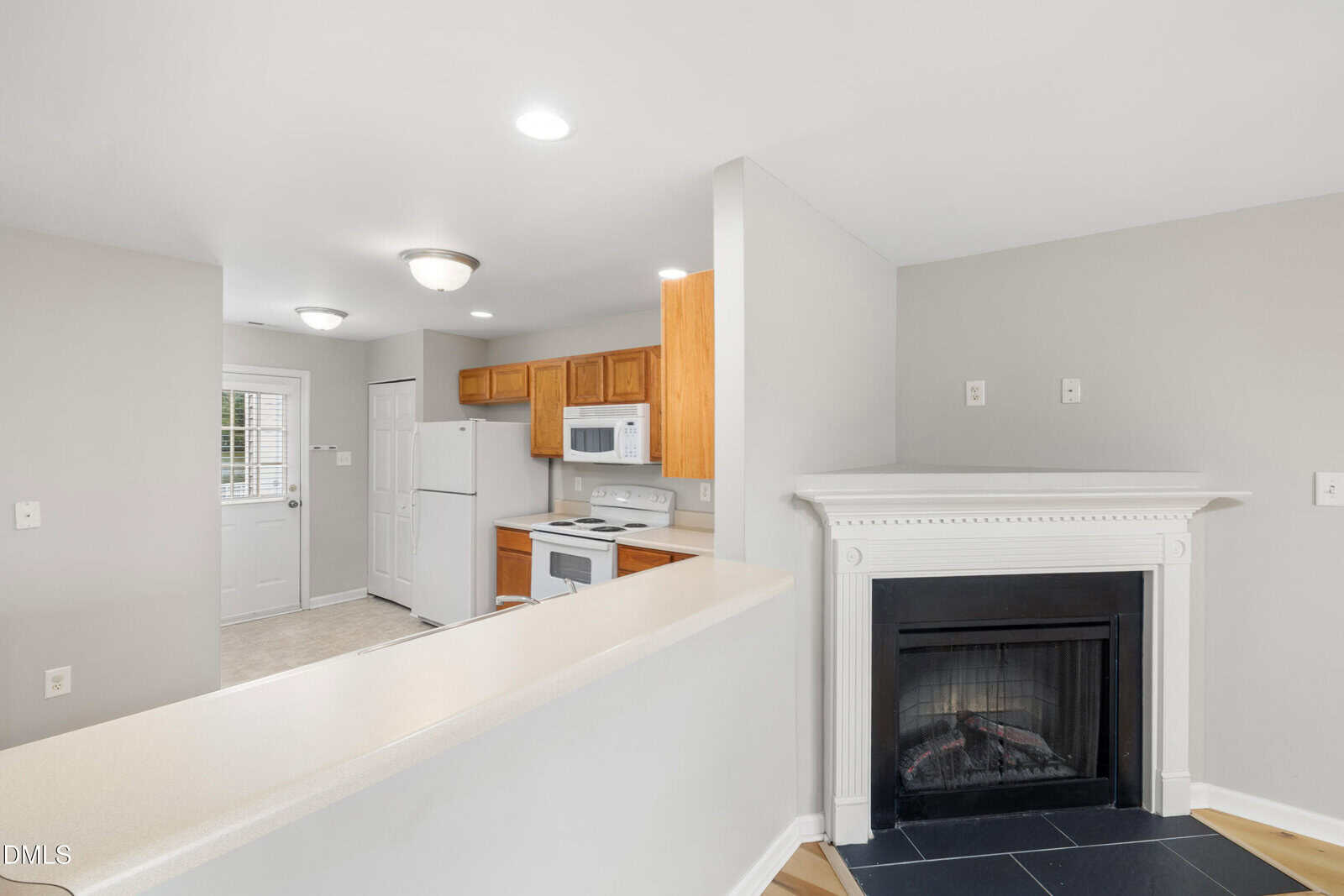 Bright kitchen with wood cabinets, white appliances, and a door leading outside. A white fireplace with a black hearth is in the foreground, and the walls are painted light gray. Ceiling lights illuminate the space.