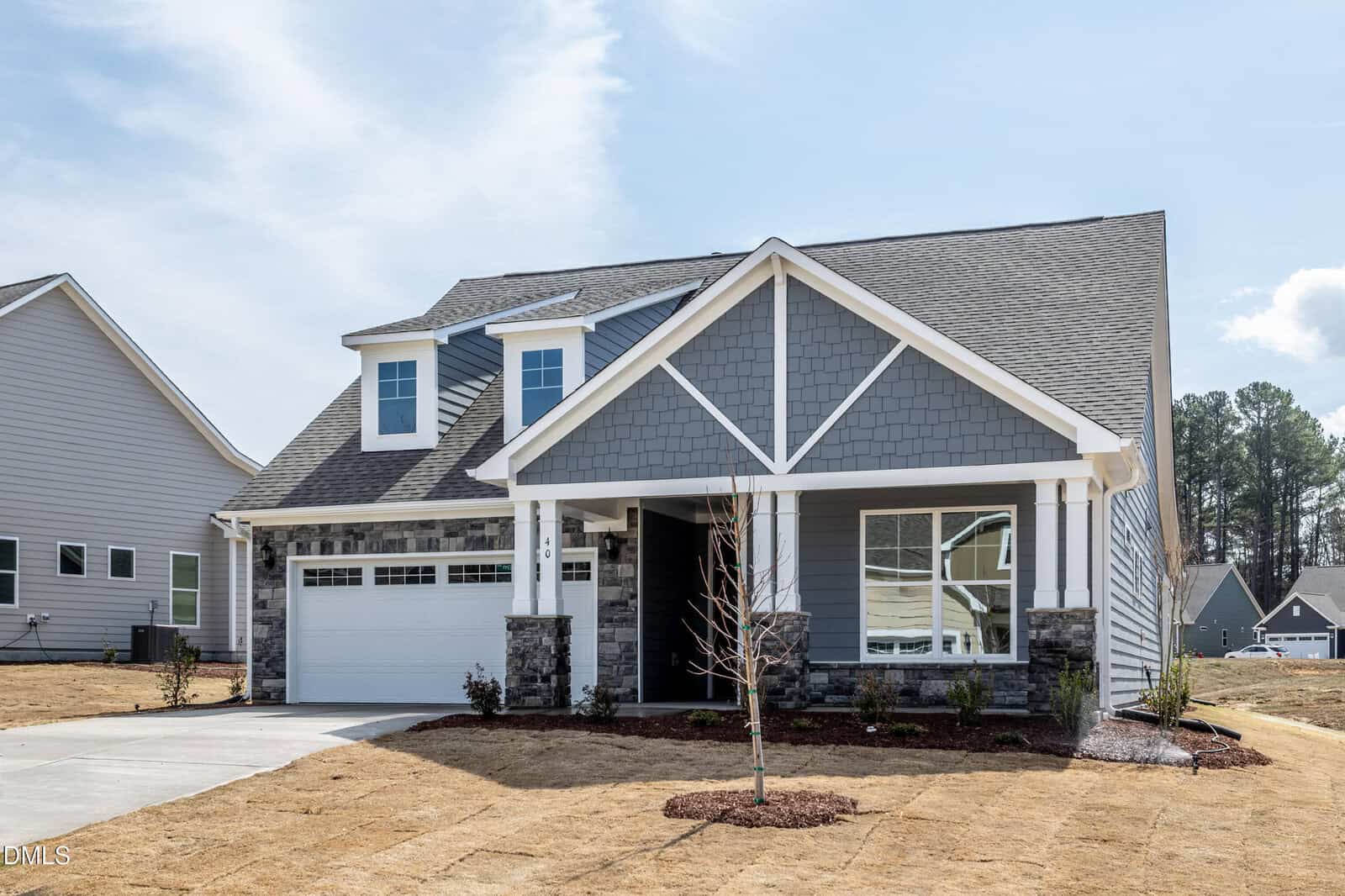 A newly built, gray two-story suburban house with white trim, a covered front porch, attached garage, and a neatly landscaped front yard under a blue sky.