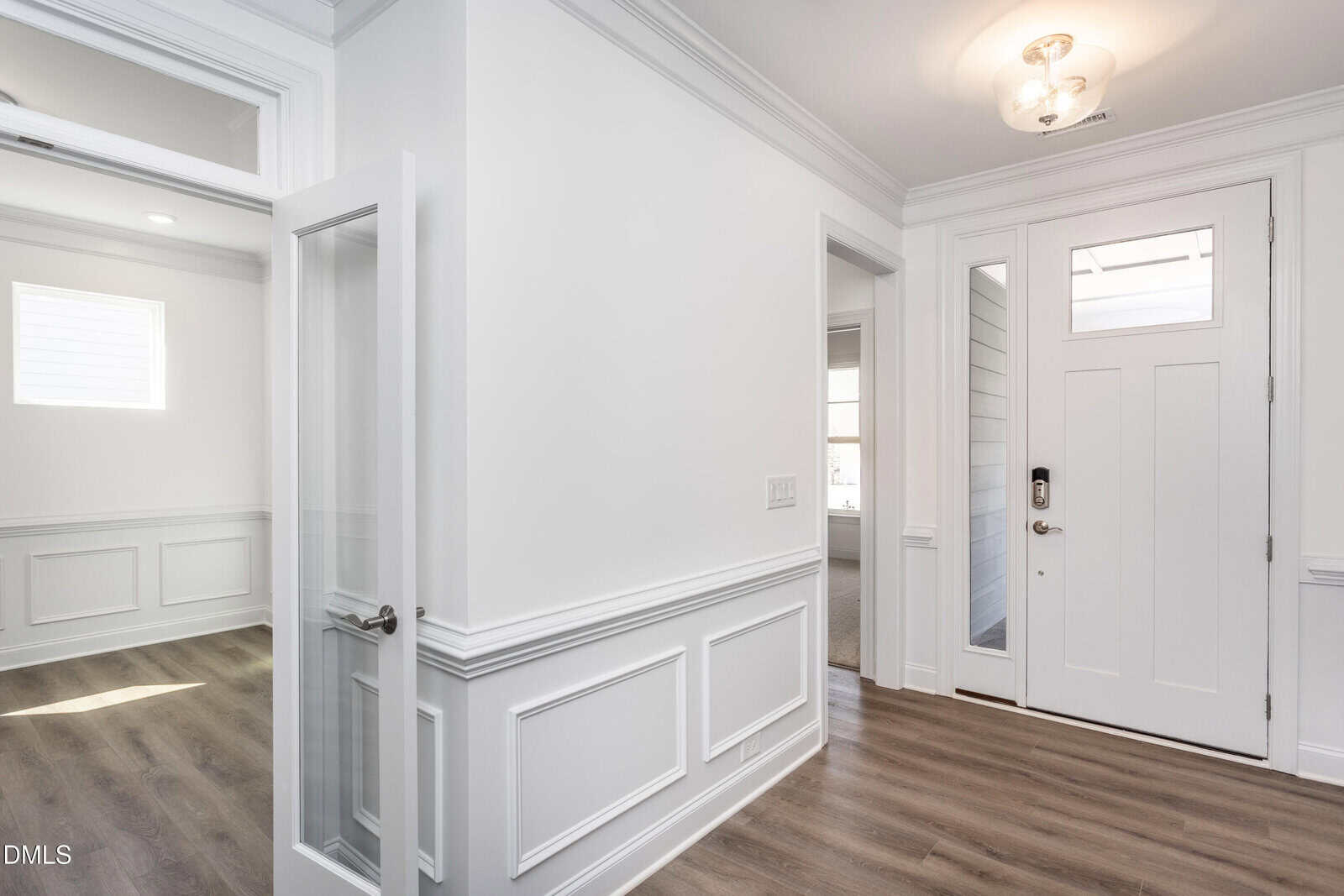 Bright entryway of a modern home featuring white walls with wainscoting, wood flooring, a glass-paneled interior door, a front door with a window, and natural light streaming through several windows.