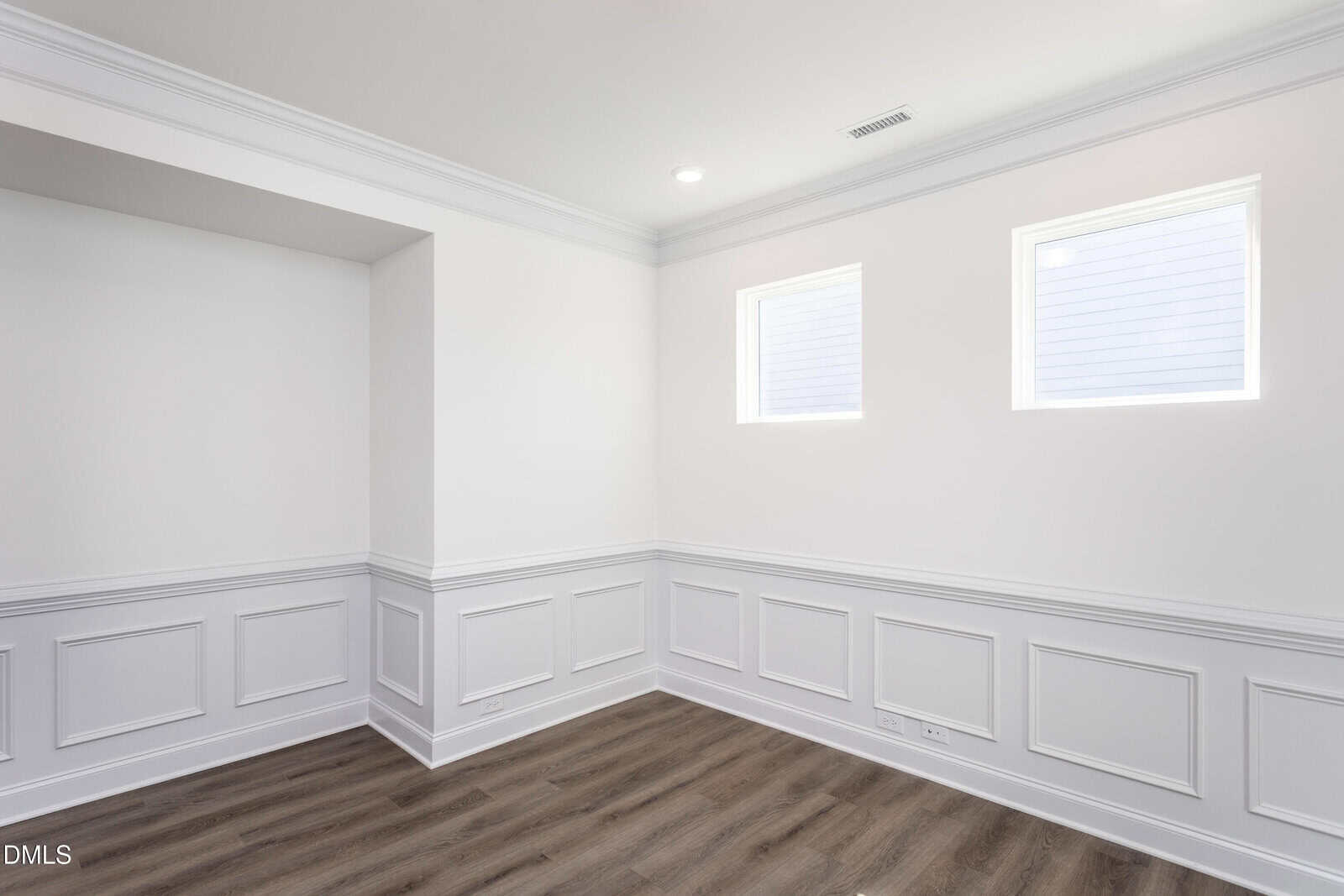 A bright, empty room with white walls, decorative wainscoting, two square windows, crown molding, and wood-style flooring. Natural light enters through the windows, creating a clean and modern look.