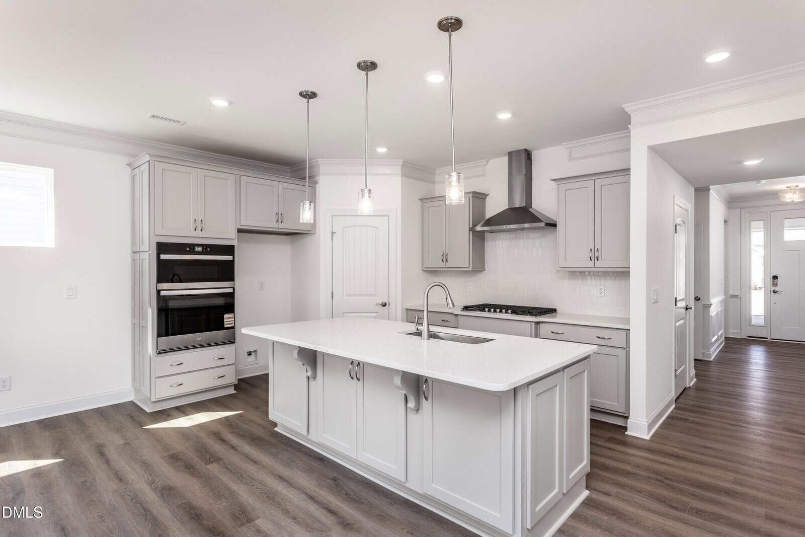 Modern kitchen with white cabinets, a large island with sink, stainless steel appliances, pendant lights, wood flooring, and an open layout leading to a hallway and front door.