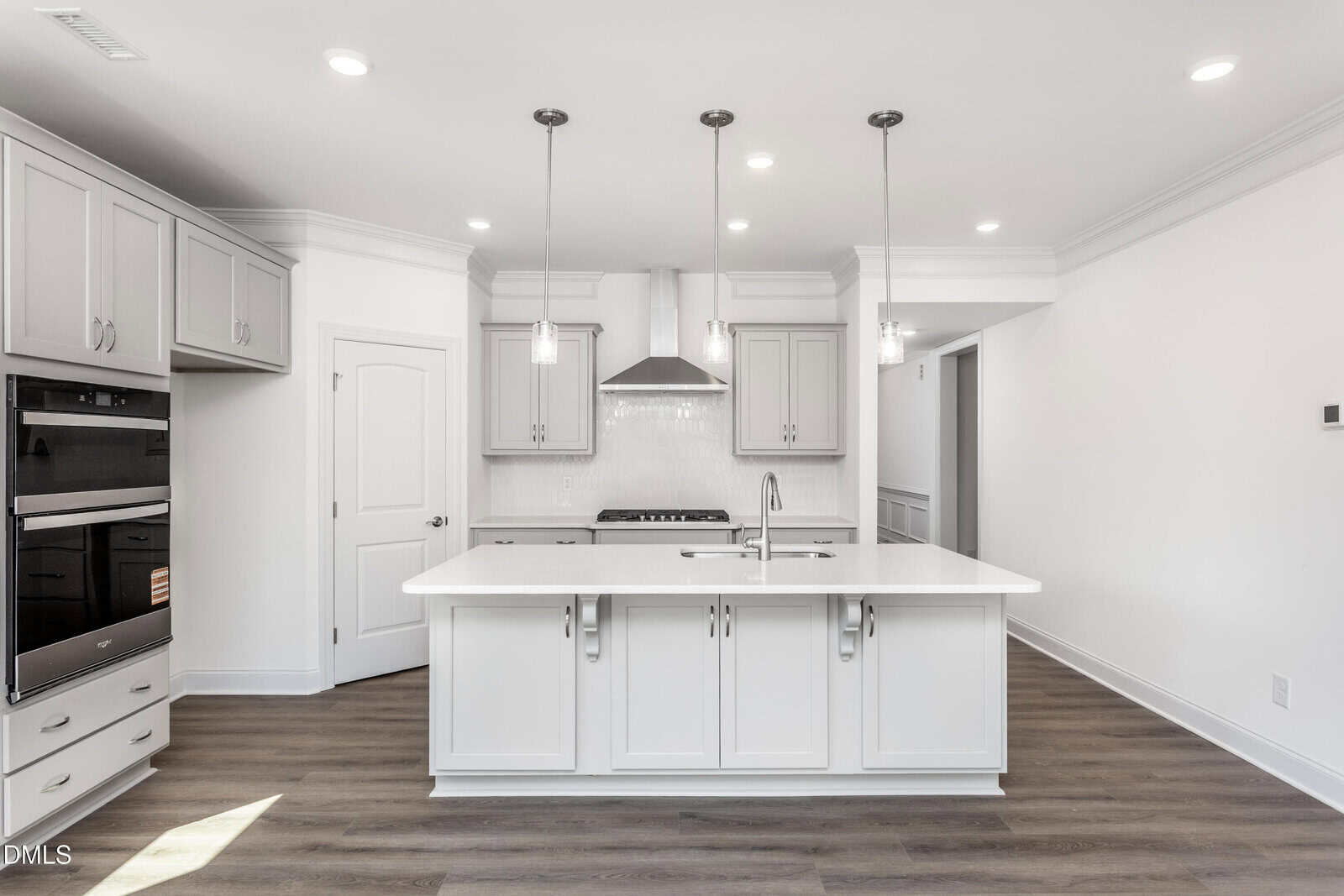Modern kitchen with white cabinets, a center island with sink, stainless steel appliances, pendant lighting, wood floor, and neutral walls. A double oven is built into the cabinetry on the left side.