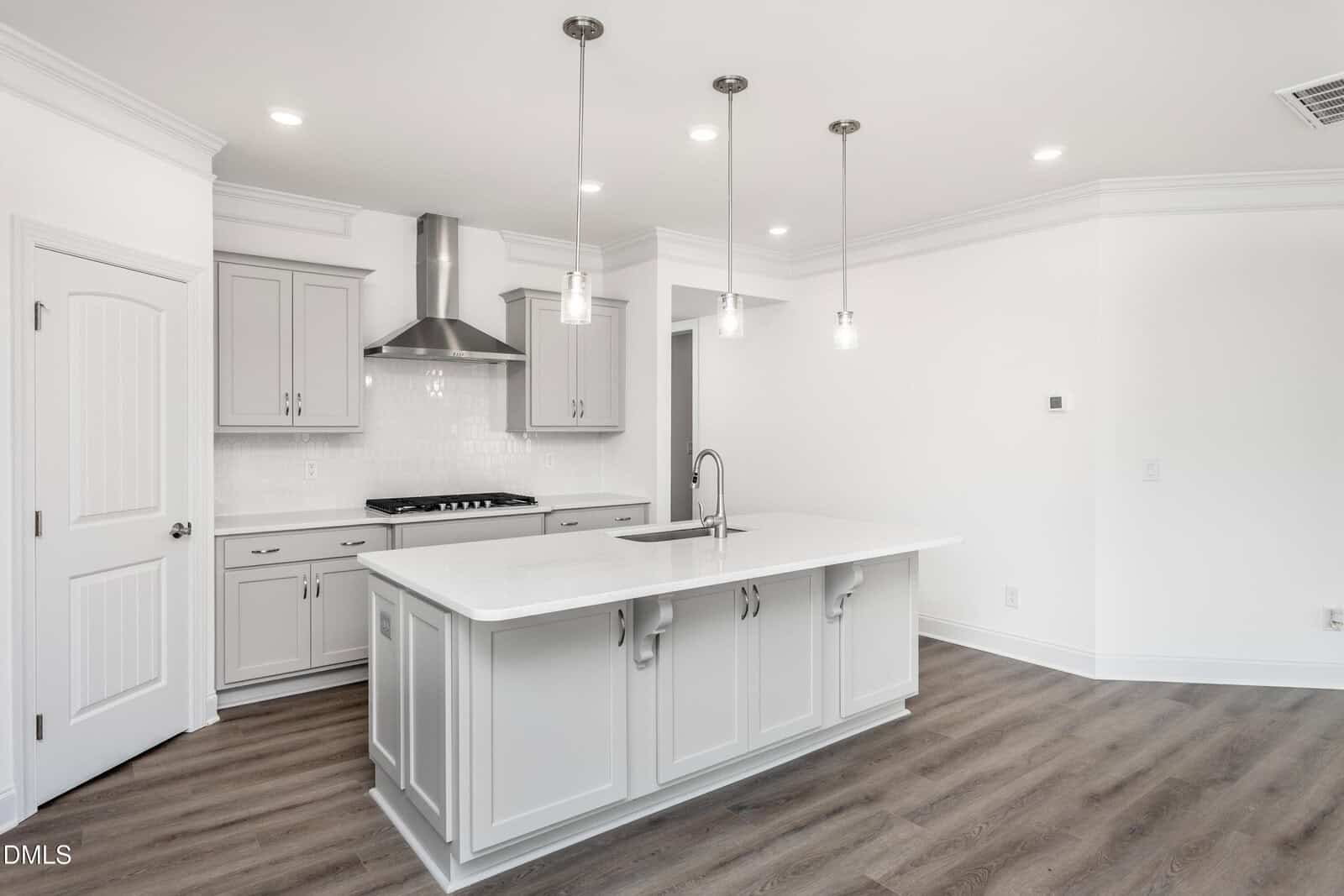 Modern kitchen with gray cabinets, white countertops, a large island with sink, stainless steel stove and hood, pendant lights, and wood-look flooring. Walls are white with crown molding and recessed lighting.