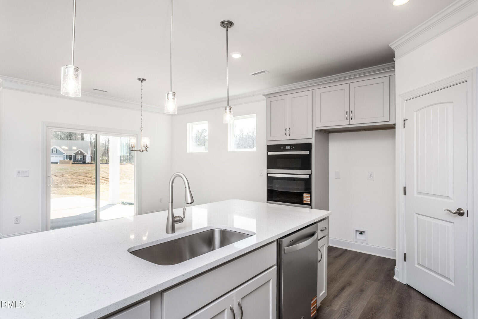 Modern kitchen with a large white island, built-in stainless steel appliances, pendant lights, light gray cabinets, a sink, and dishwasher. Natural light enters from windows and a glass door leading outside.