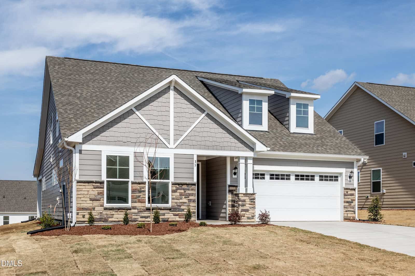 Single-story modern suburban house with gray siding, white trim, stone accents, and a two-car garage. Fresh landscaping surrounds the home, and the sky is clear and blue.