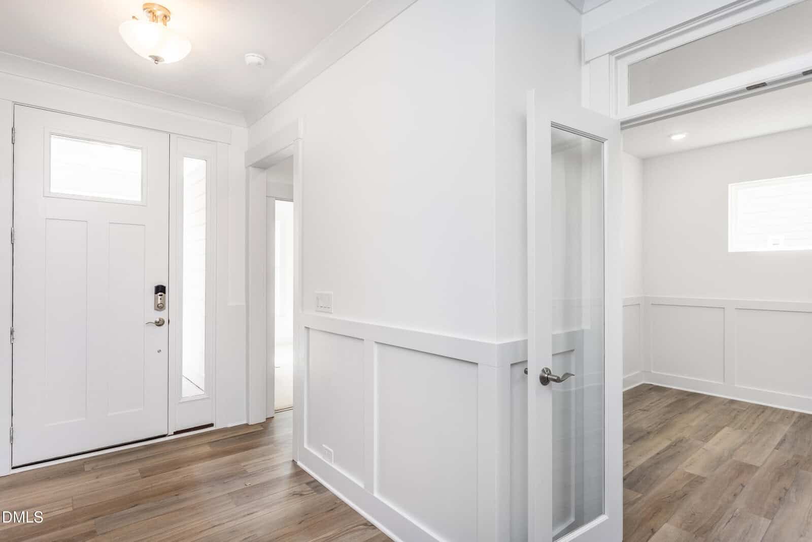 Bright entryway with white walls, trims, and door, wood flooring, and a glass-paneled room entrance on the right. Natural light comes through windows beside and above the front door.
