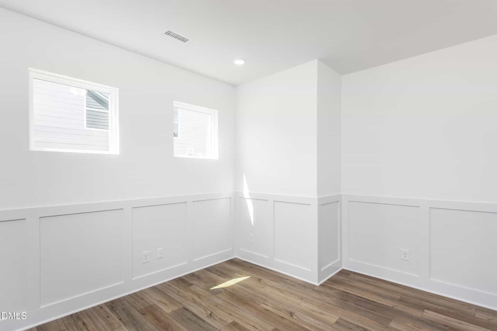 Bright, empty room with white walls, white wainscoting, two small windows, recessed lighting, and light wood flooring. The room has a clean, modern look with natural light streaming in.