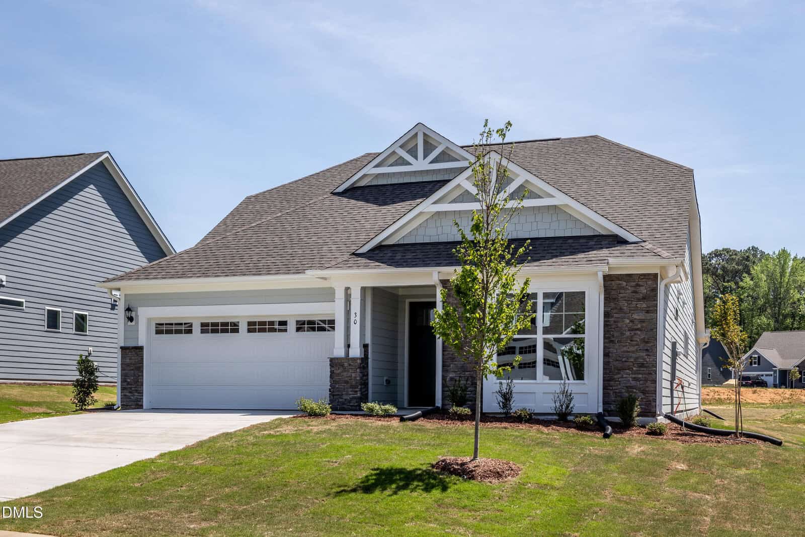A modern single-story house with gray siding, white trim, a two-car garage, and a small front yard with young trees and fresh landscaping on a sunny day.