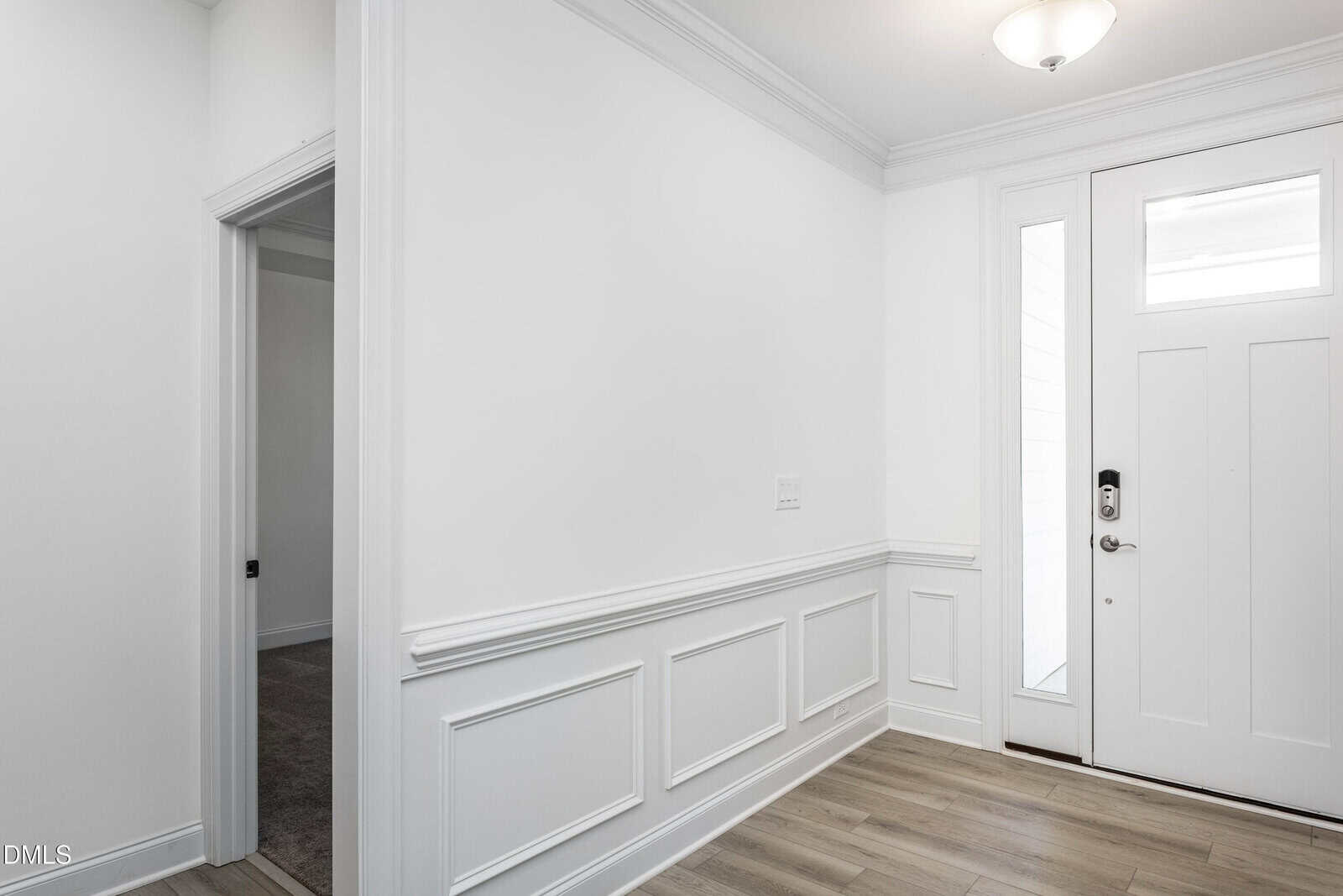 A bright, modern entryway with white walls, decorative wainscoting, wood flooring, and a white front door with windows. A doorway on the left leads to another room.