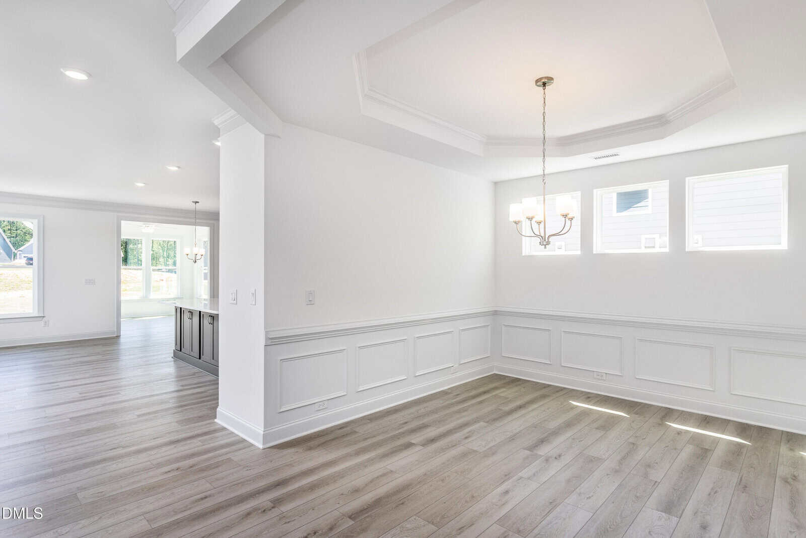 Bright, empty dining room with white walls, coffered ceiling, chandelier, and large windows. Light wood floors extend into an adjacent open living area with more windows and natural light.