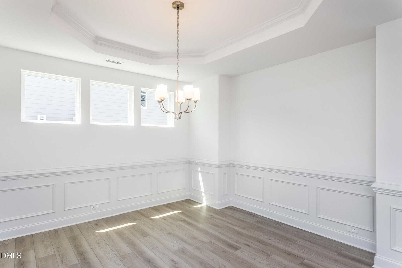 Bright, empty dining room with three high windows, white walls with wainscoting, light wood floors, and a modern chandelier hanging from a tray ceiling. Daylight streams in, creating a clean, airy feel.