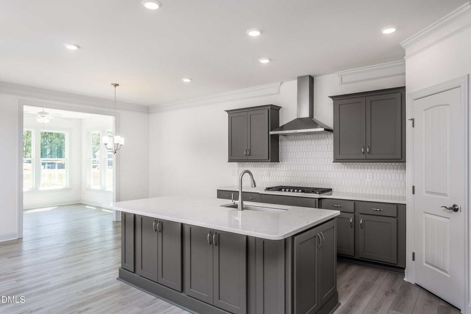 Modern kitchen with gray cabinets, a large island with a sink, stainless steel appliances, white tile backsplash, and bright dining area with large windows in the background. Light wood floors throughout.
