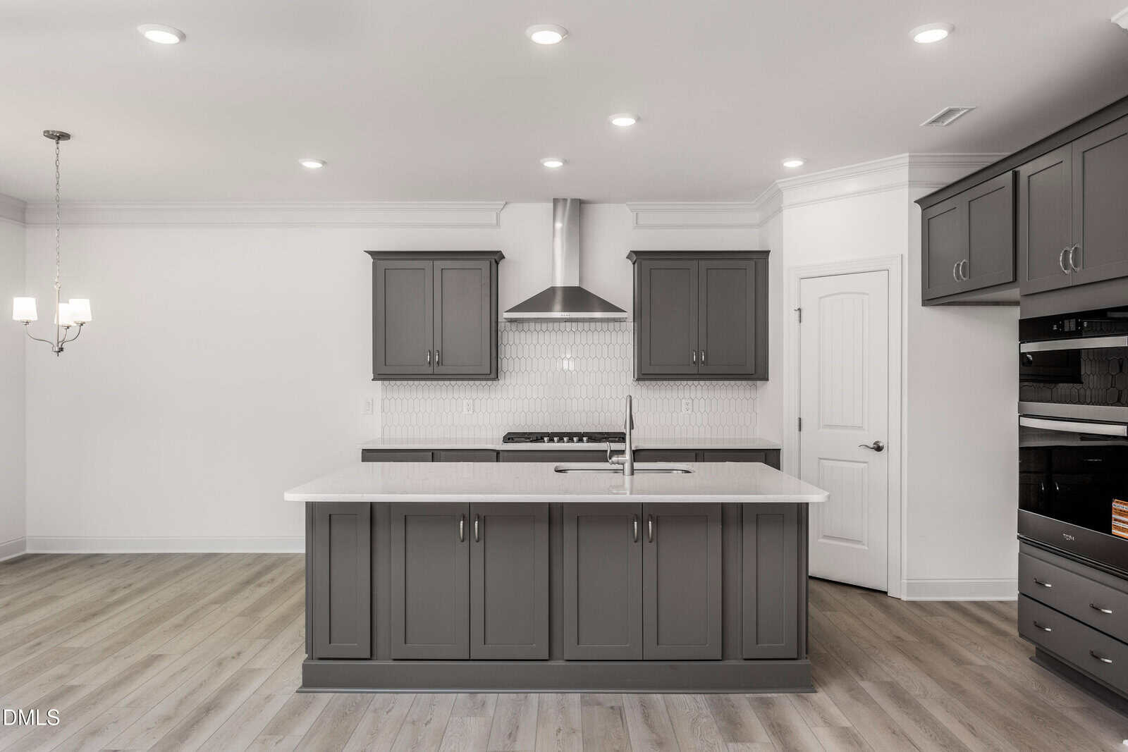 Modern kitchen with gray cabinets, white subway tile backsplash, stainless steel appliances, an island with a sink, light wood flooring, and a chandelier in the adjacent dining area. The space has recessed ceiling lights.