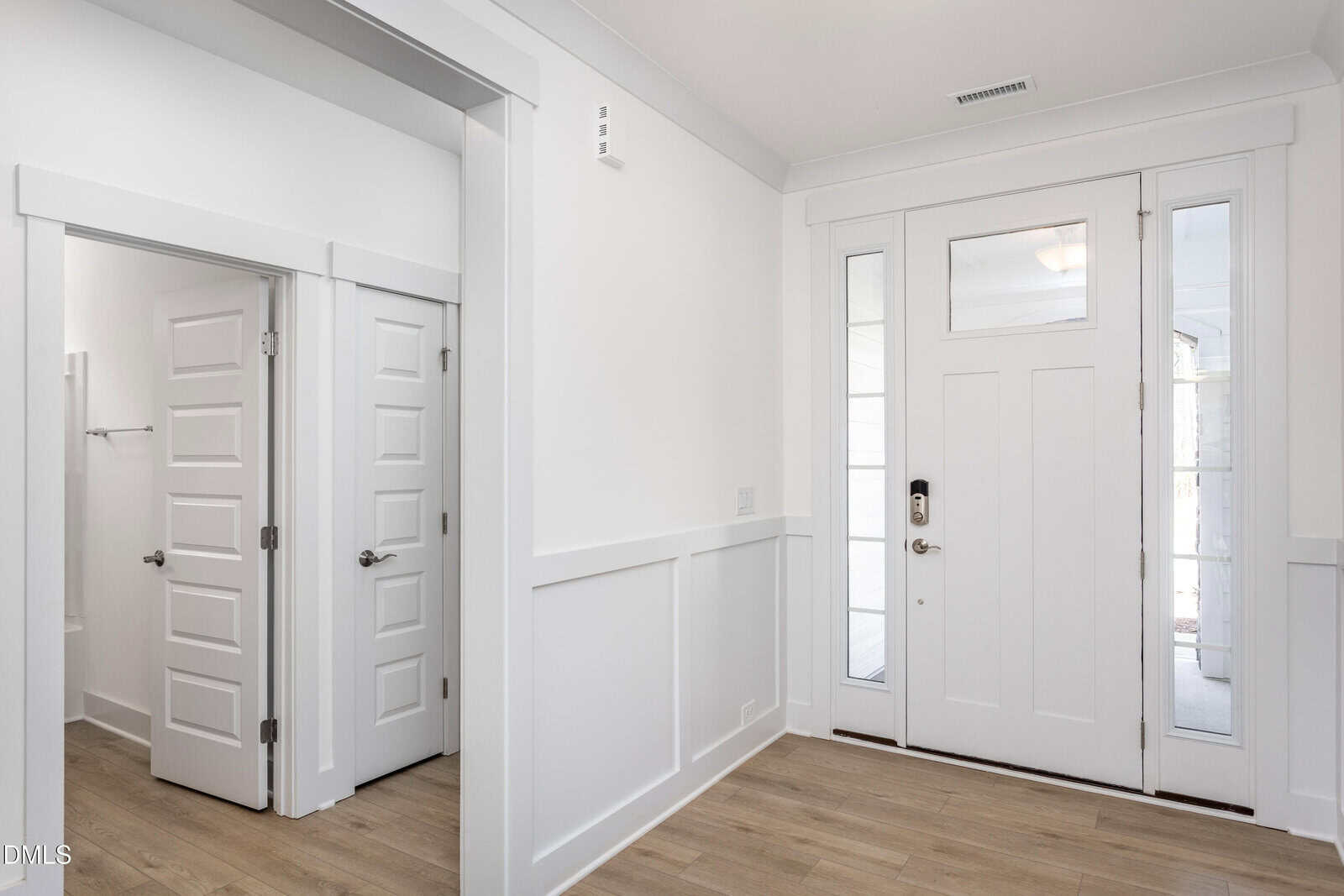 Bright entryway with light wood flooring, white walls, and a white front door with glass panels. Two white interior doors, one open and one closed, are visible to the left.