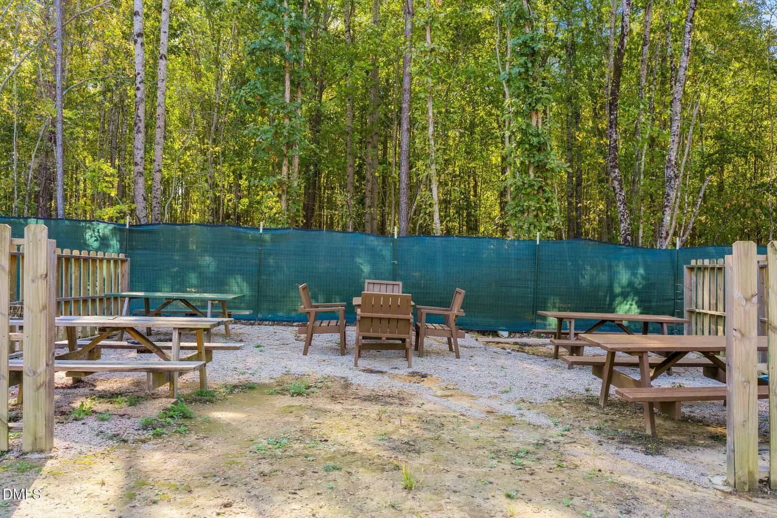Several wooden picnic tables and chairs are arranged outdoors on gravel, enclosed by a wooden fence and a green mesh barrier. Tall trees line the background. The area is quiet and shaded.